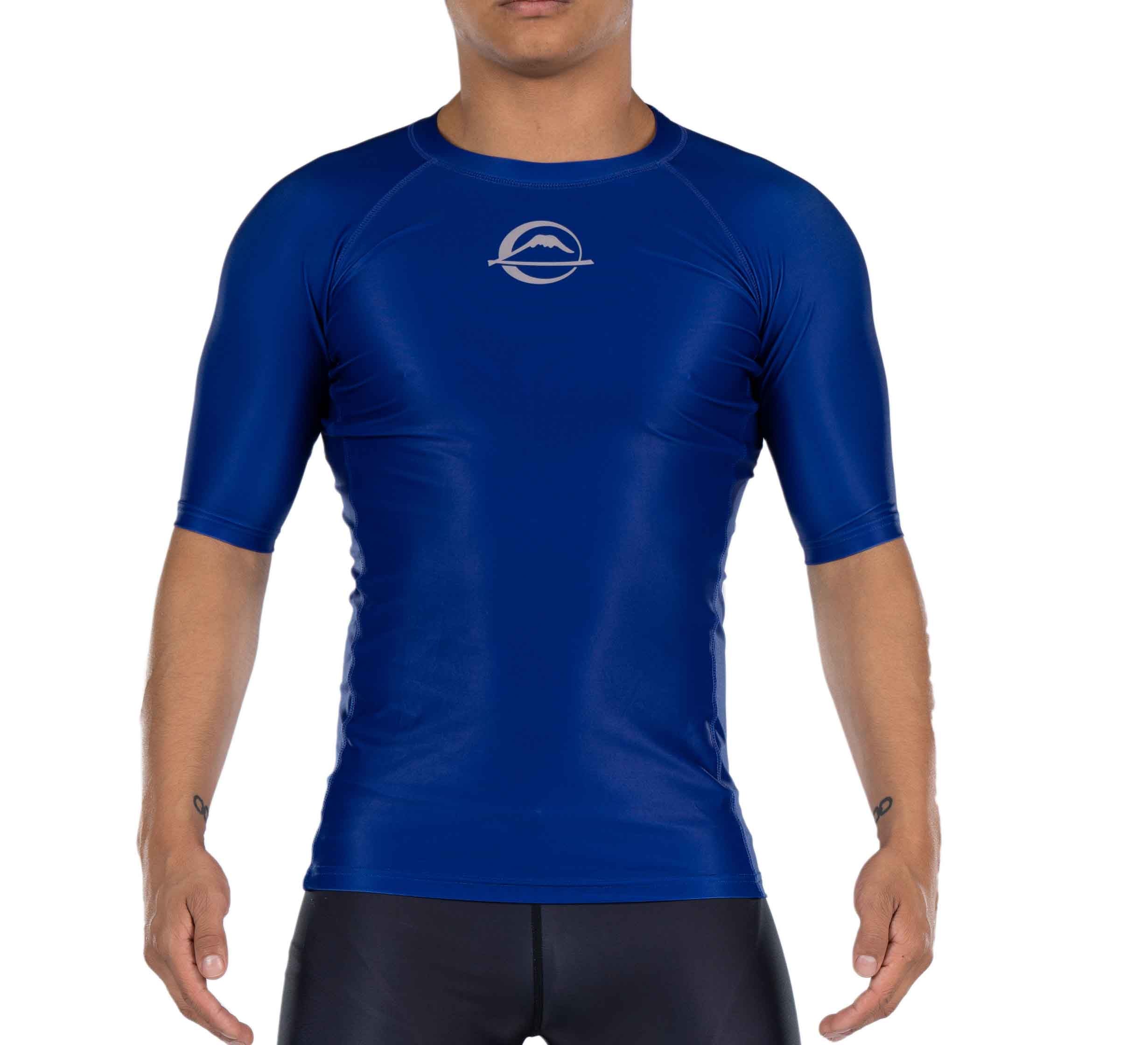 A person wearing the Baseline Ranked Short Sleeve Rashguard Blue, featuring a fitted cut, short sleeves, and a white circular chest logo, stands with arms slightly away from their sides against a plain background.