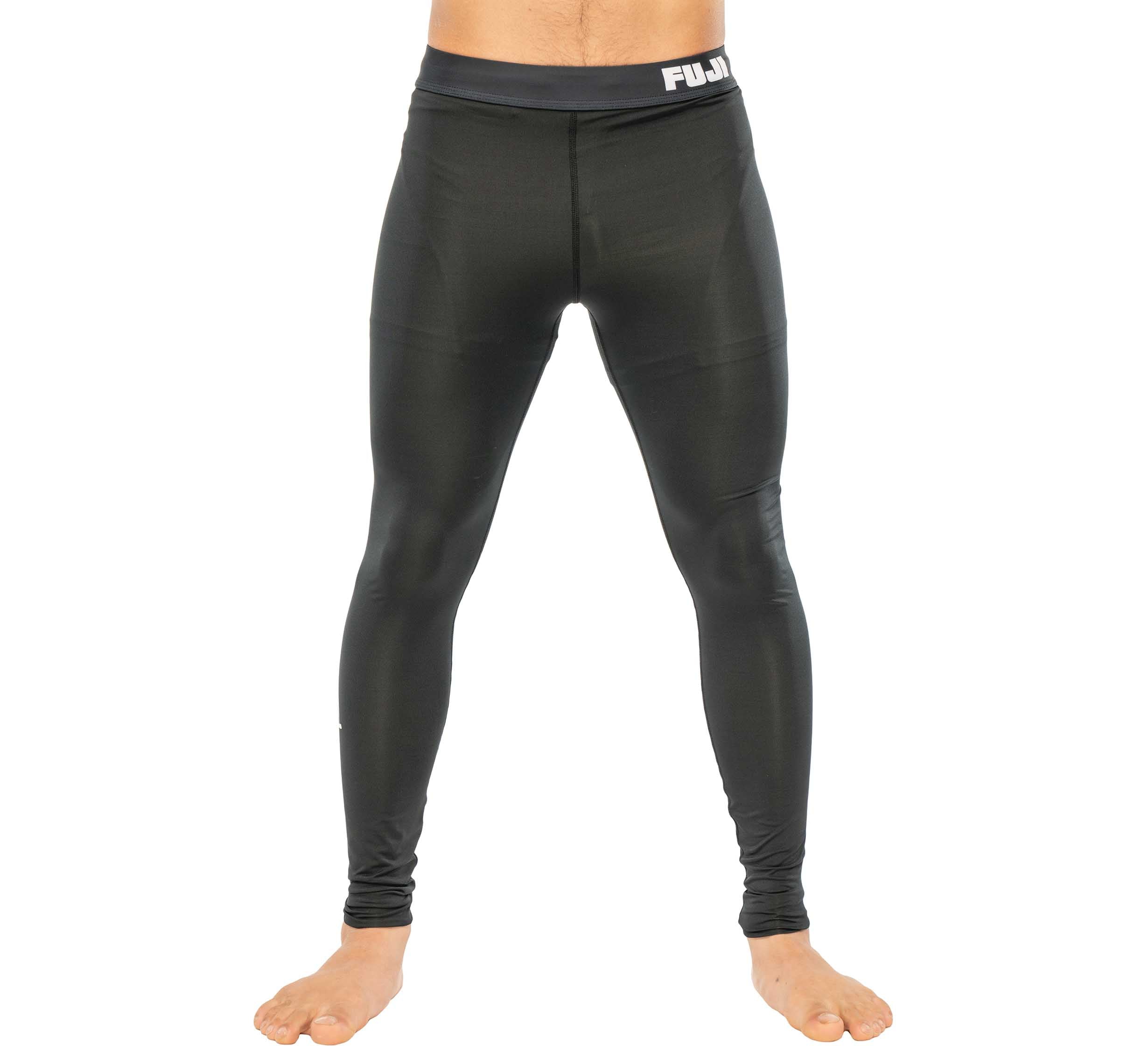 A person stands barefoot against a plain white background, wearing Baseline Grappling Spats V2 black athletic compression leggings with a partially visible FUJI logo on the waistband. The upper body is not shown.