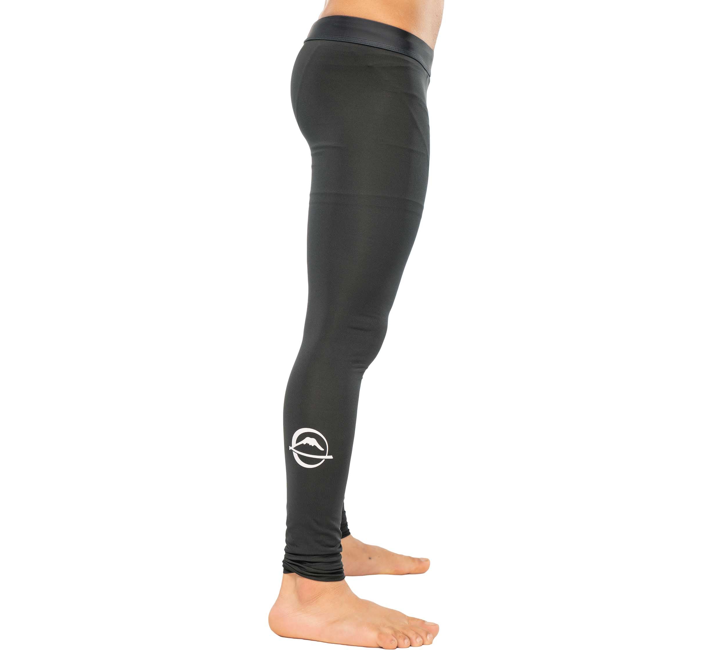 A person stands barefoot in Baseline Grappling Spats V2, black fitted leggings featuring a white circular logo near the ankle, shown side profile against a plain white background.