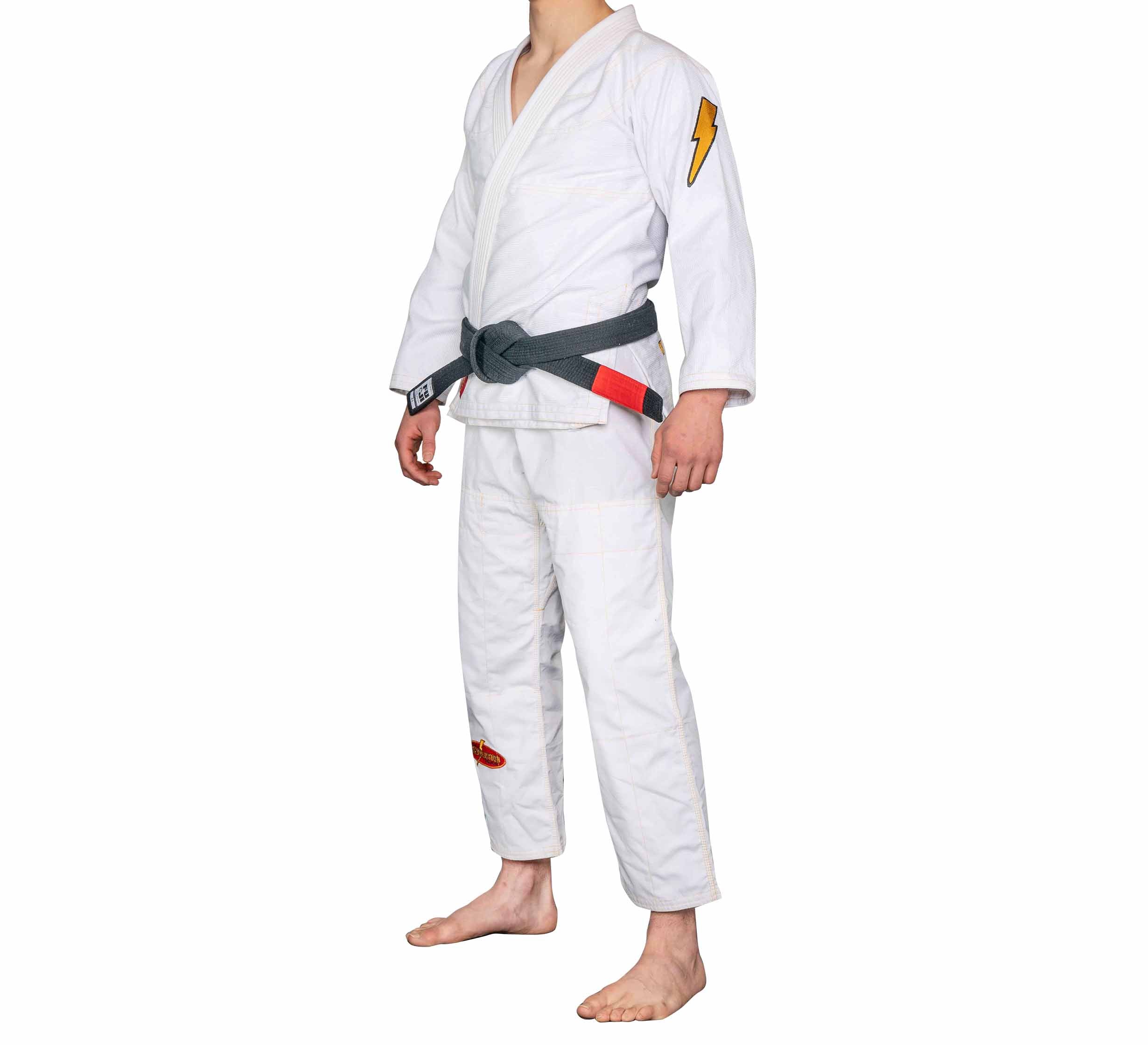 Person wearing the BJJ Revolution Warrior Gi with a gray belt, barefoot on a white background. The gi features a yellow lightning bolt patch on the left sleeve and a small red patch on the right pant leg.