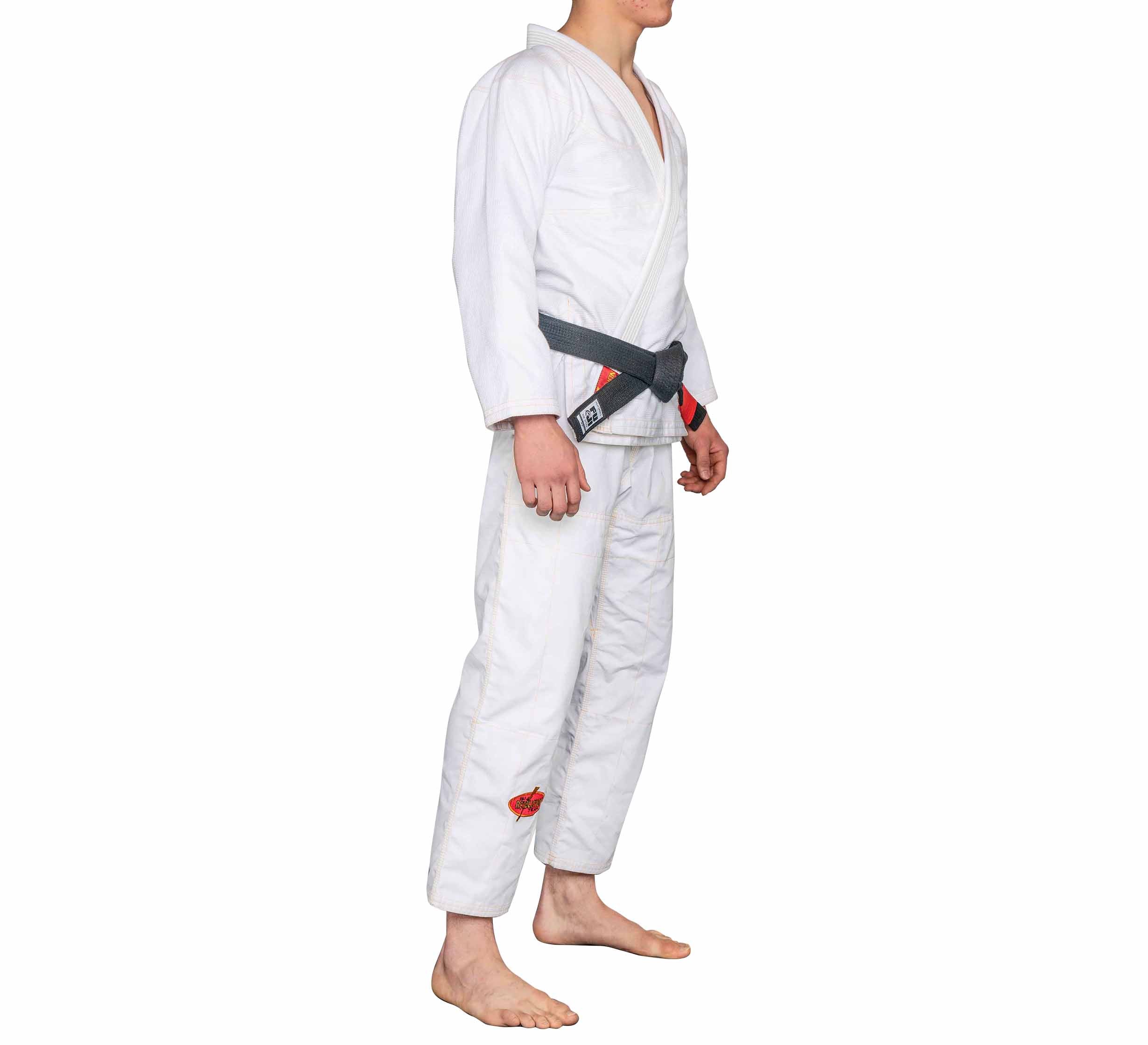 A person wearing the BJJ Revolution Warrior Gi with a black belt stands barefoot on a white background, shown from the side with arms relaxed at their sides.