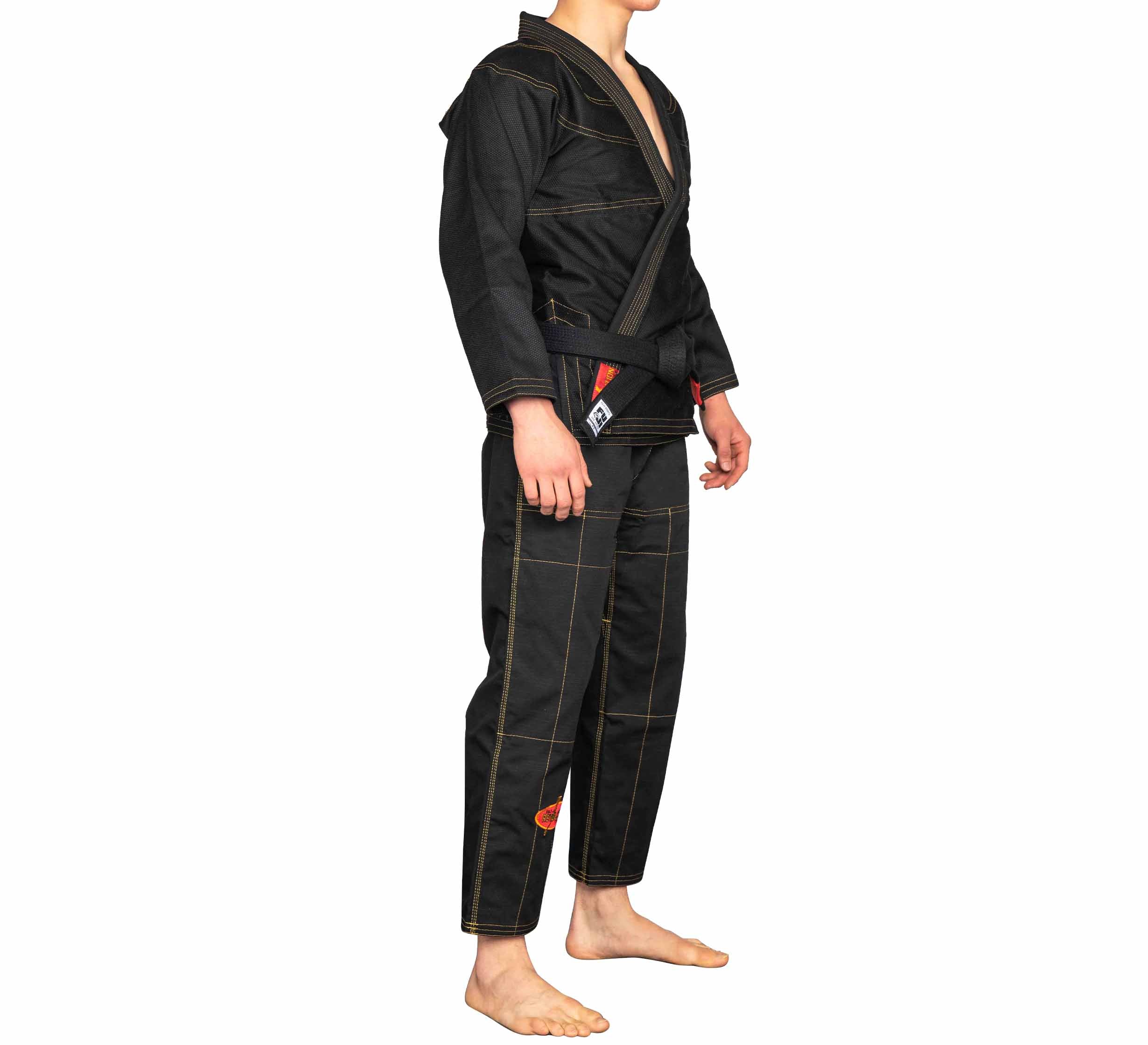A person stands barefoot on a plain white background, wearing the BJJ Revolution Warrior Gi—black with visible stitching, orange accents, and finished with a black belt.