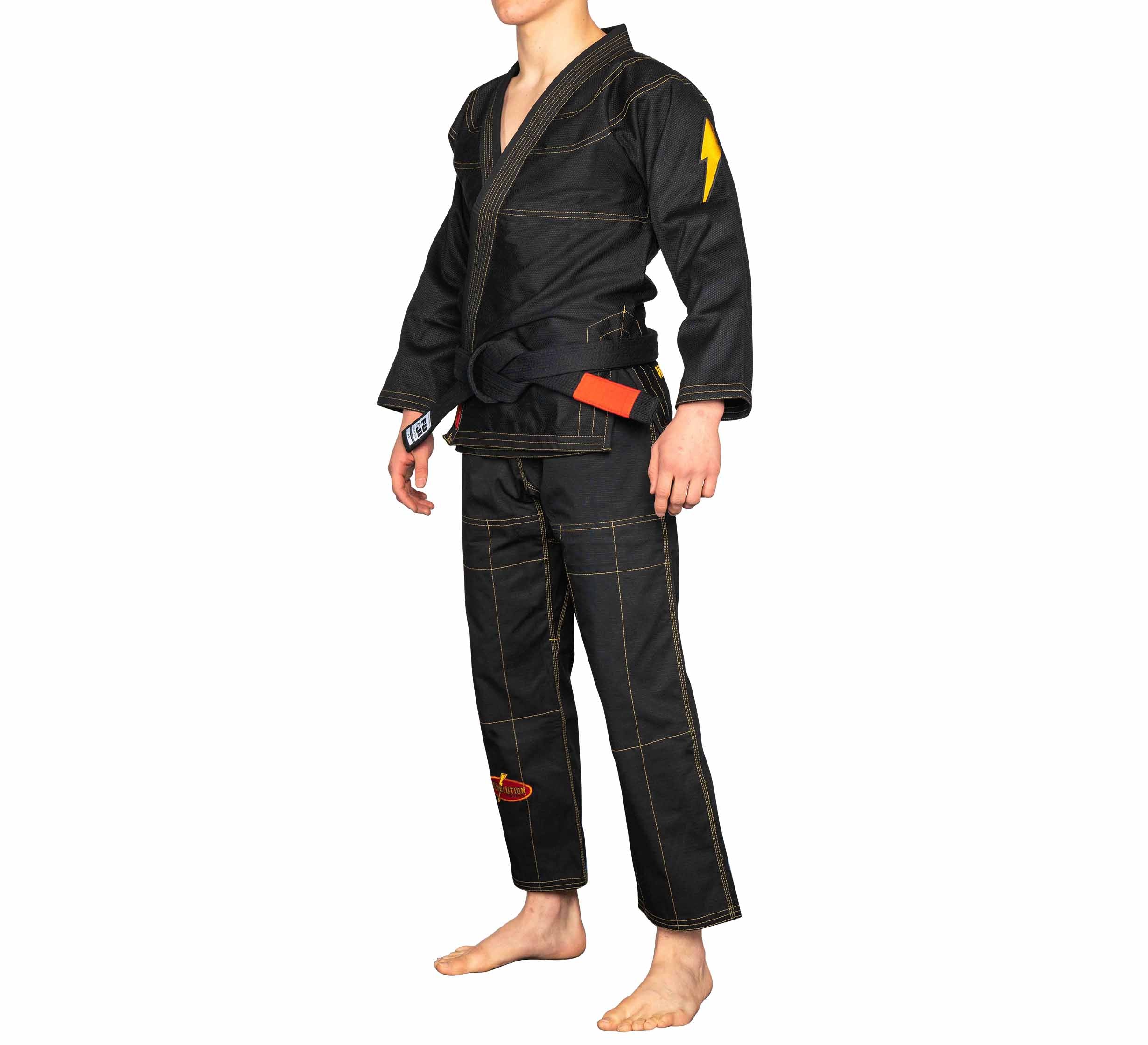 A person wears the BJJ Revolution Warrior Gi, a black martial arts uniform with contrast stitching, a black belt, and a yellow lightning bolt patch on the left sleeve, standing barefoot against a plain white background.