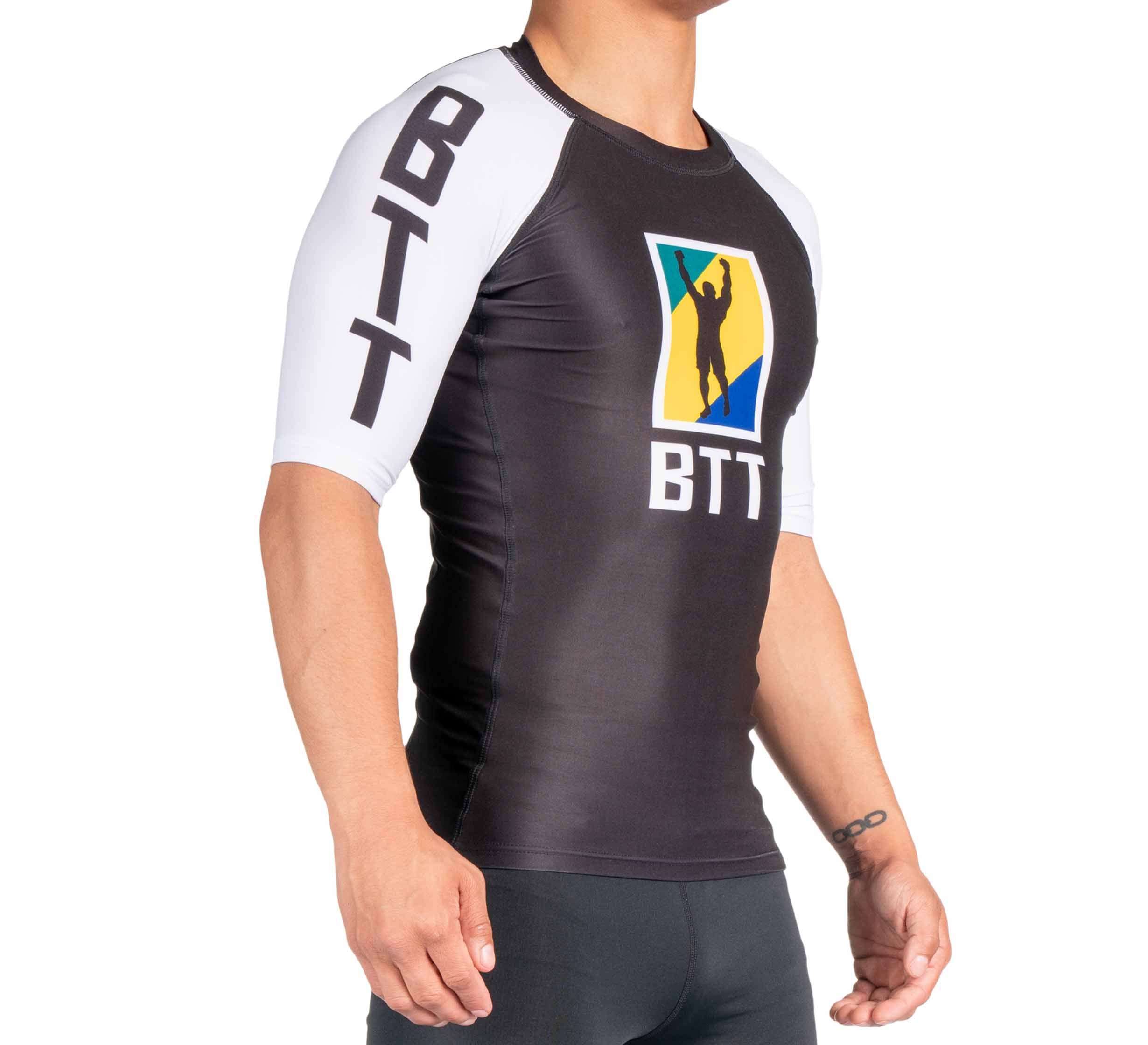 A person wearing the BTT Ranked Short Sleeve Rashguard (NEW), featuring black and white colors, BTT on the sleeves and chest, plus a colorful shield logo with a silhouette of a person raising one arm.