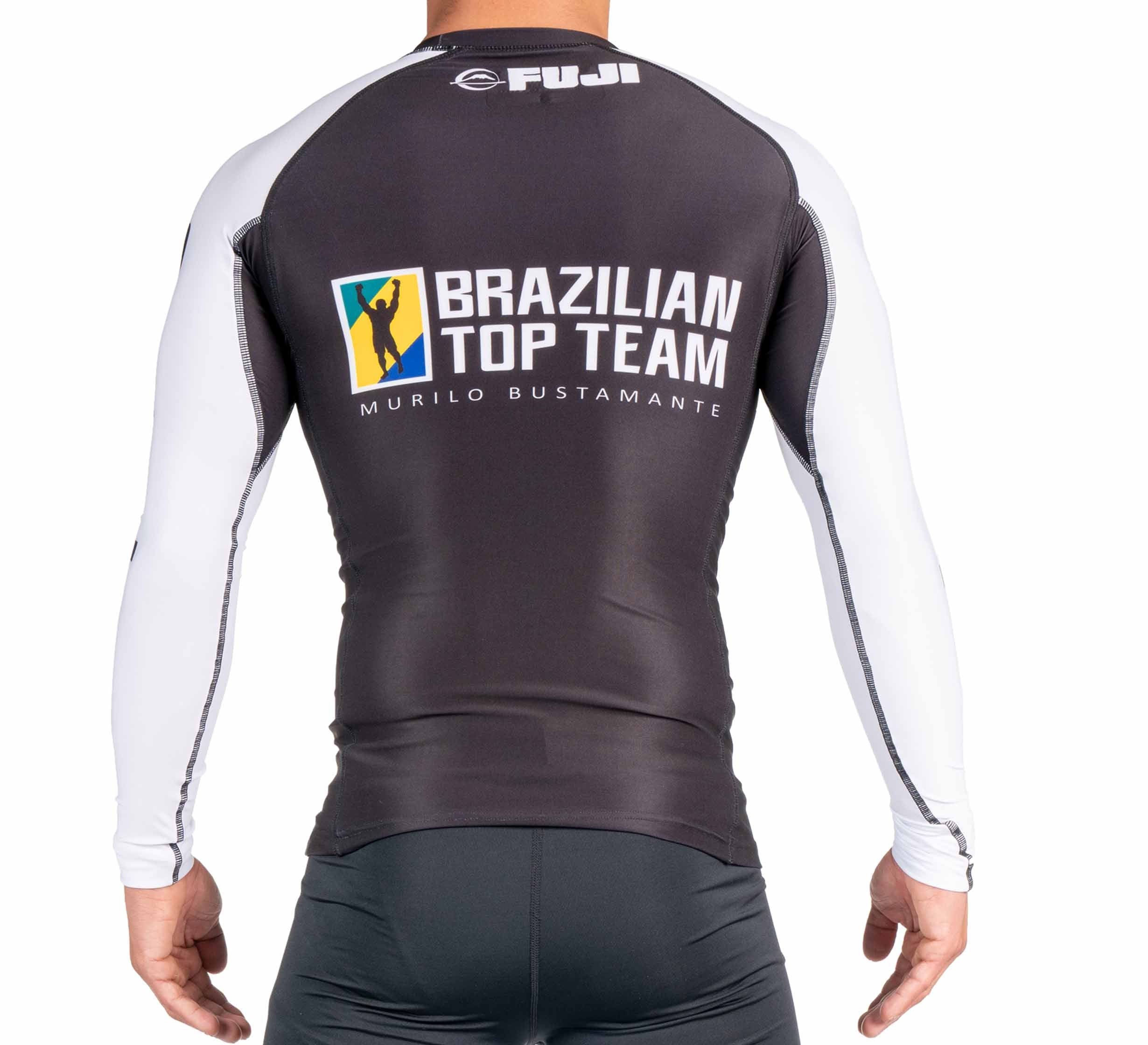 A person wears the BTT Ranked Long Sleeve Rashguard (NEW), black and white with long sleeves. The back displays the FUJI logo and a large Brazilian Top Team Murilo Bustamante logo featuring a yellow and blue graphic.