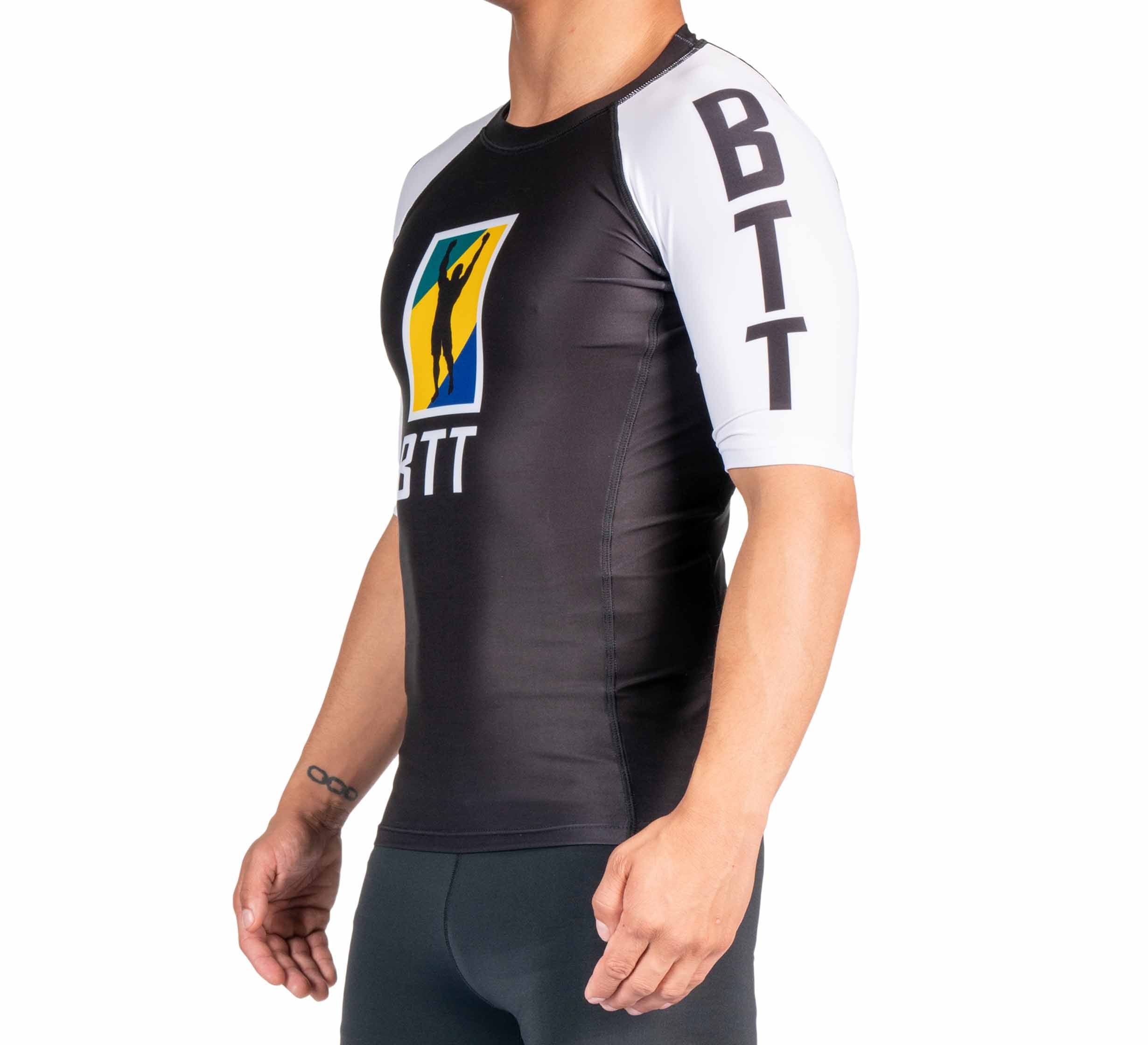 A person in the BTT Ranked Short Sleeve Rashguard (NEW), featuring black and white colors, BTT on the sleeve, and a colorful chest logo, stands in side profile against a white background.