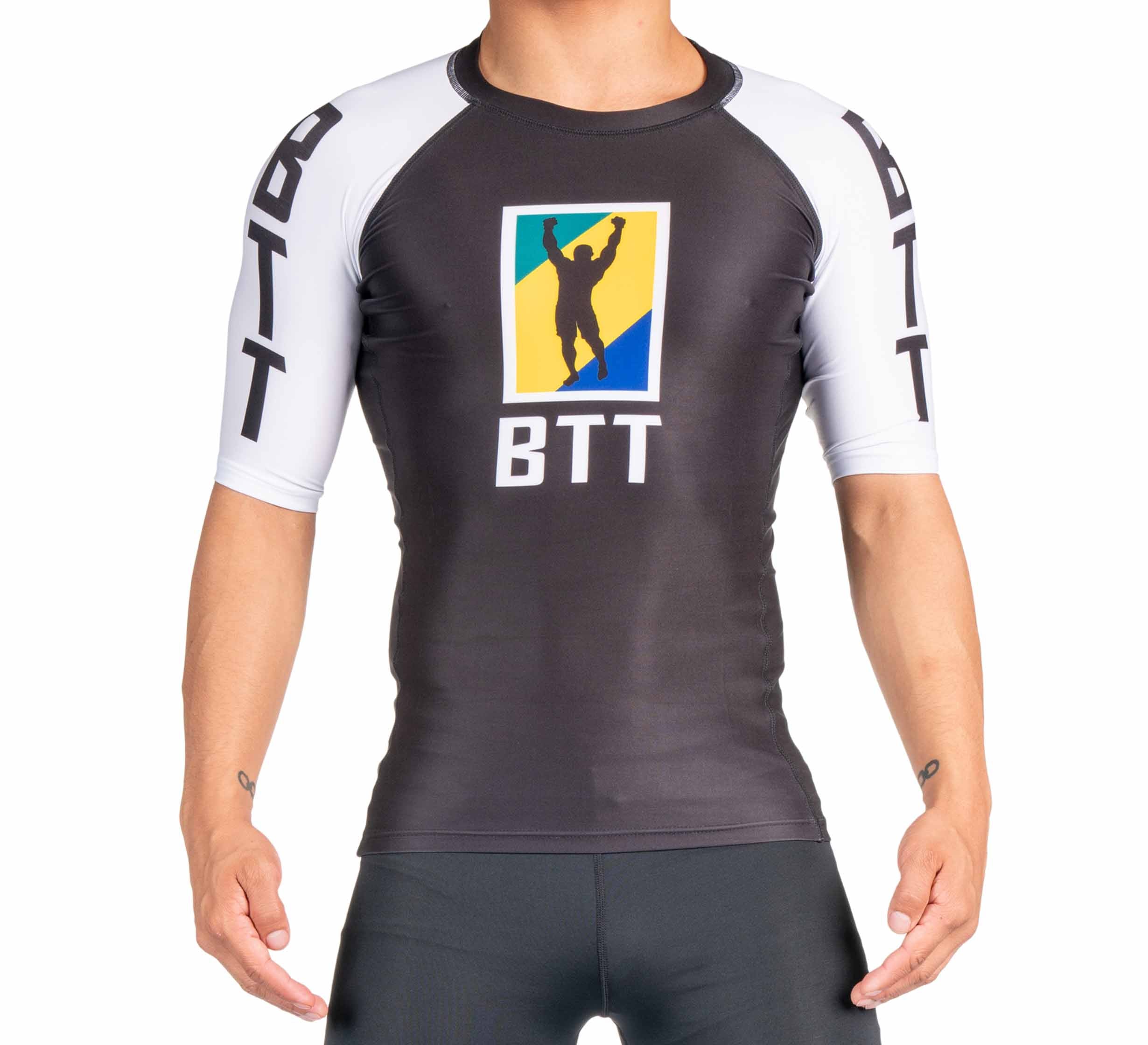 A person wears the BTT Ranked Short Sleeve Rashguard (NEW), a black and white athletic top with “BTT” text on the chest and sleeves, plus a colorful triumphant figure logo displayed on the chest.