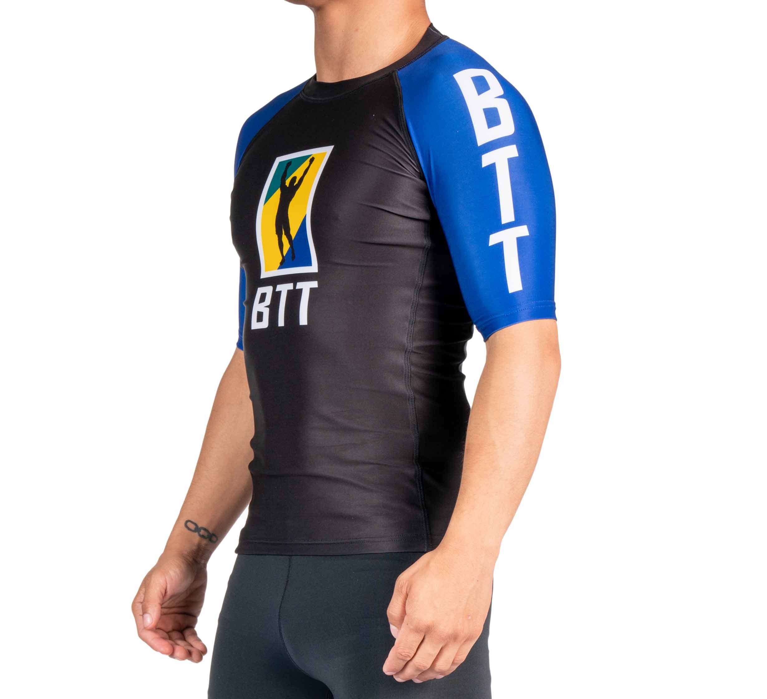 A man stands sideways against a plain white background wearing the BTT Ranked Short Sleeve Rashguard (NEW) in black and blue, featuring BTT lettering and a colorful logo on the chest and sleeve.