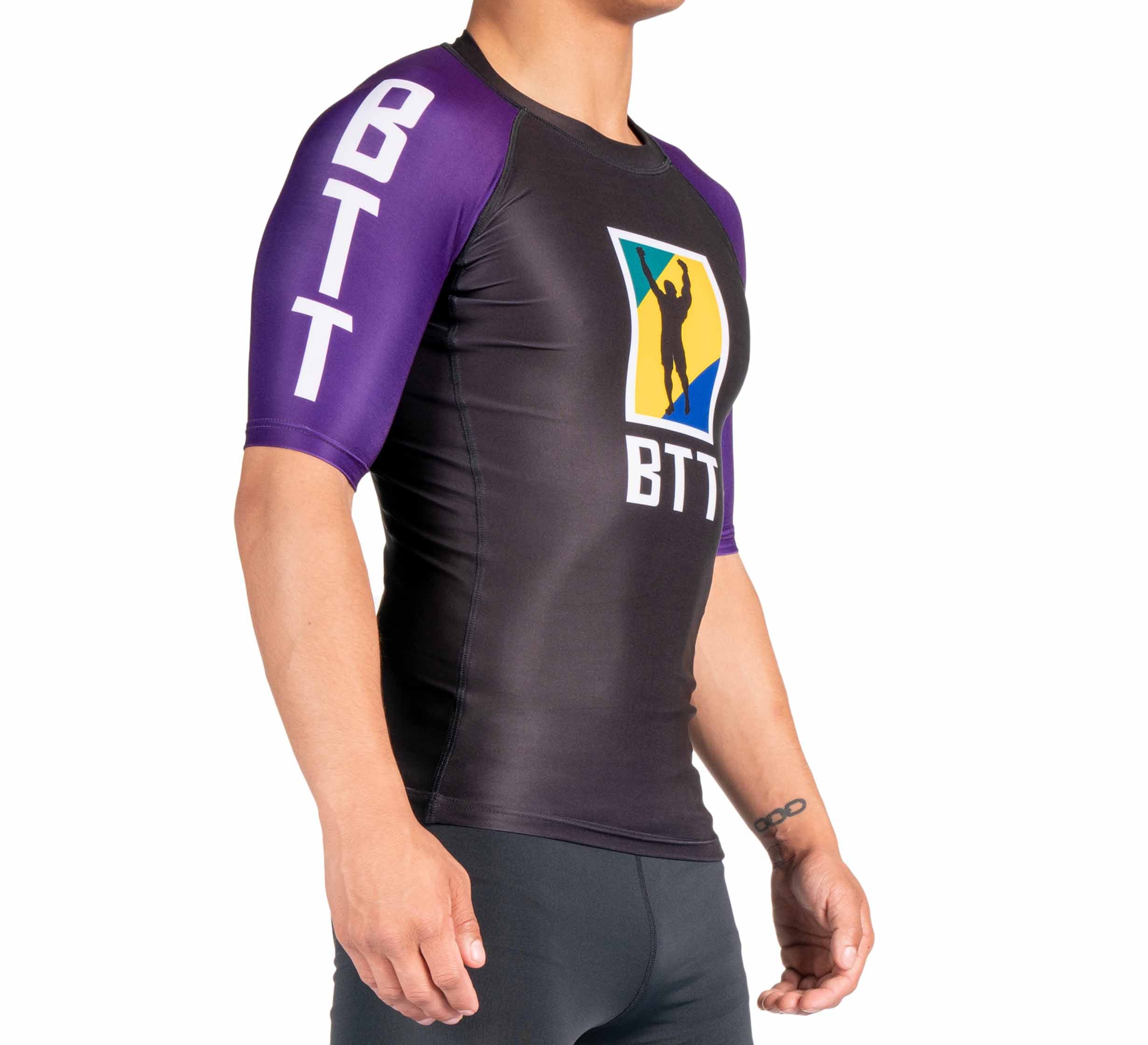 A person wears the BTT Ranked Short Sleeve Rashguard (NEW), a fitted black and purple athletic top with "BTT" on the shoulder and chest, plus a colorful logo featuring a central silhouette. The background is white.