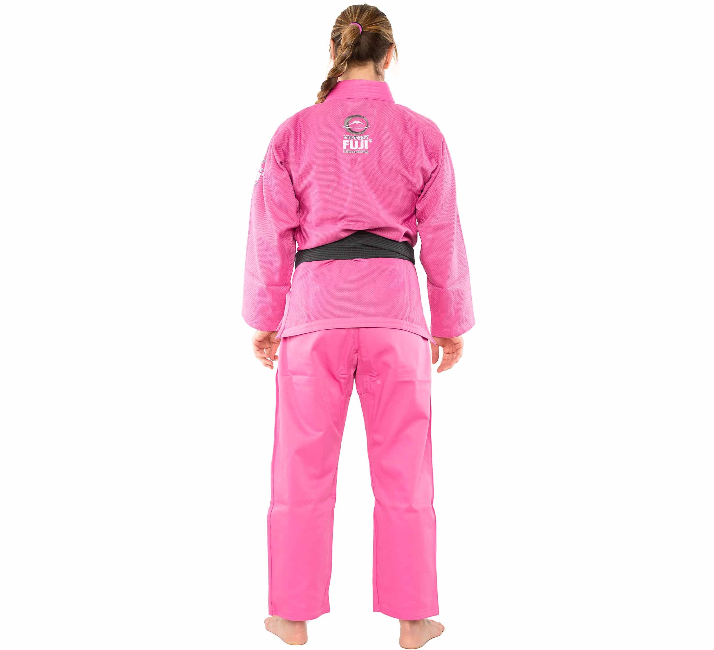 A person with braided hair stands barefoot, facing away, wearing the bright pink All Around Womens BJJ Gi by Fuji, complete with a black belt and Fuji logo on the back.