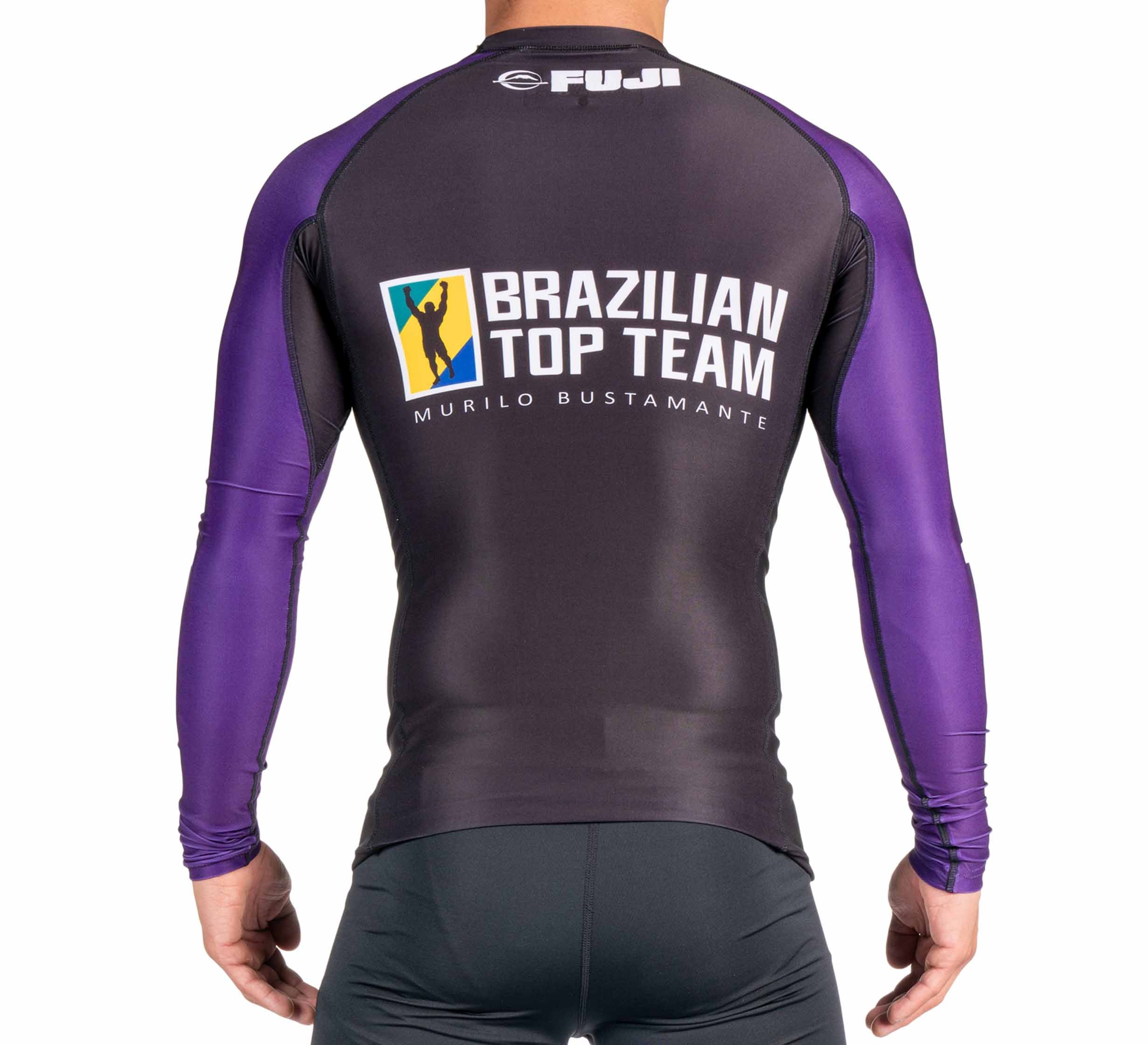 A person wearing the BTT Ranked Long Sleeve Rashguard (NEW) in black and purple, featuring Brazilian Top Team Murilo Bustamante and FUJI logos on the back, stands with their back to the camera.