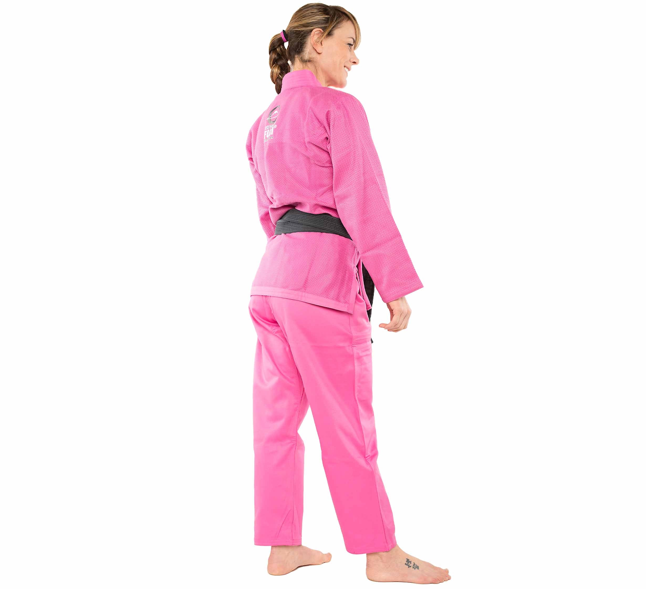 A woman wears the All Around Womens BJJ Gi Pink and a black belt, standing barefoot on a white background, facing away but smiling over her shoulder.