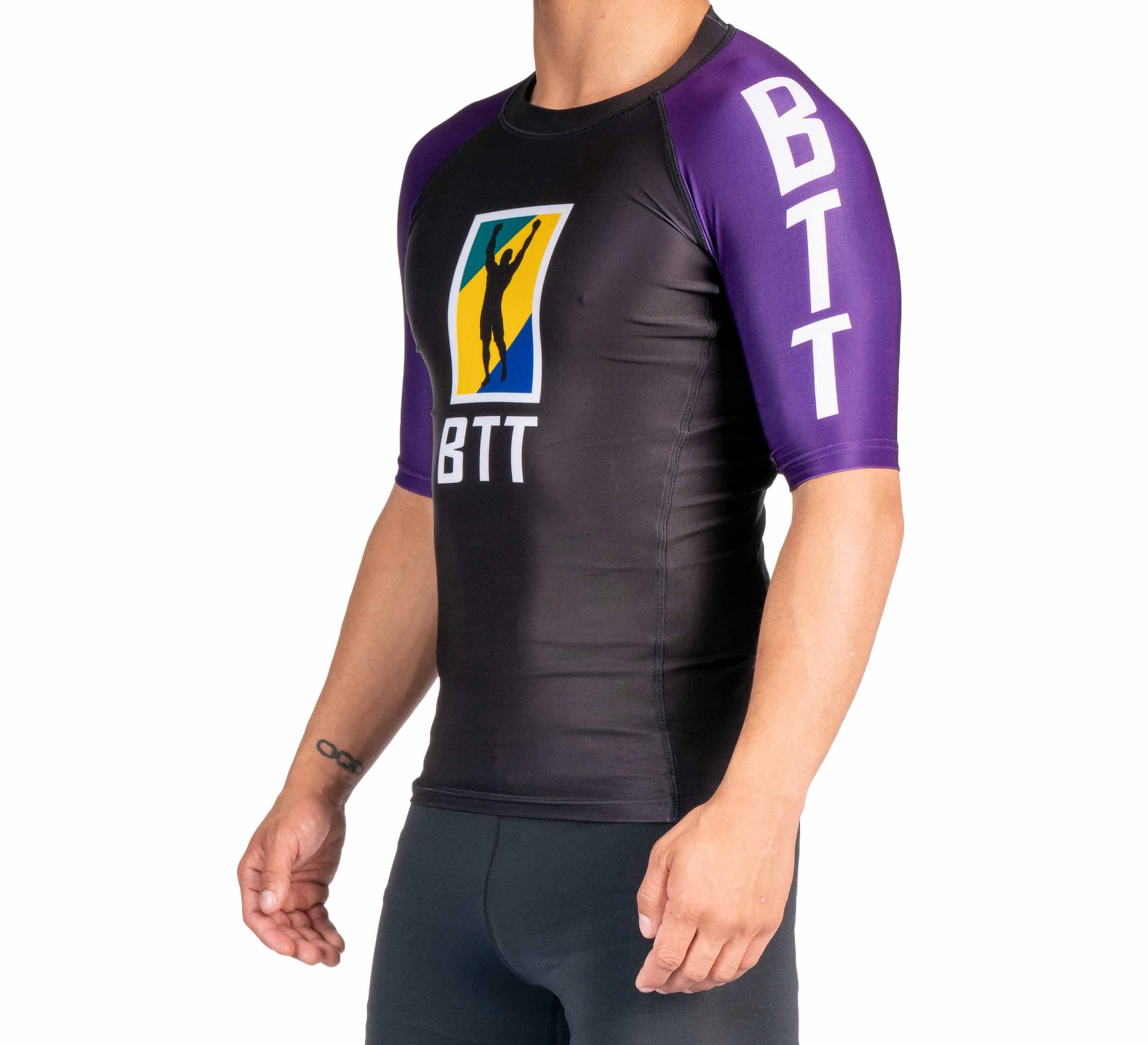 A person wearing the BTT Ranked Short Sleeve Rashguard (NEW), a fitted black and purple athletic shirt with "BTT" on the sleeve and chest and a colorful logo with a silhouette on the front.