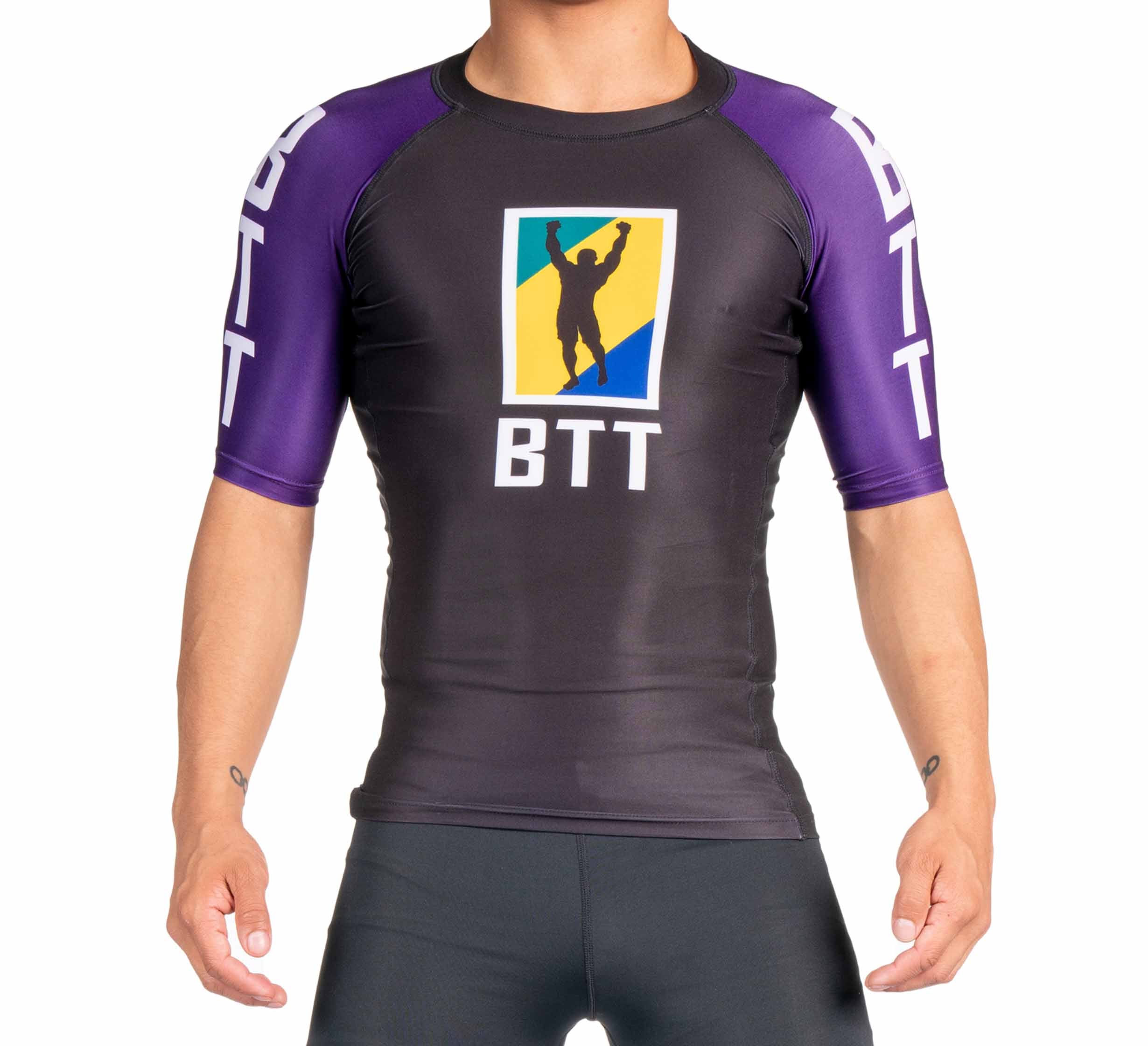 A person wearing the BTT Ranked Short Sleeve Rashguard (NEW) in black and purple, featuring the BTT logo with a figure raising their arms before a yellow and blue background. The person's face is not visible.