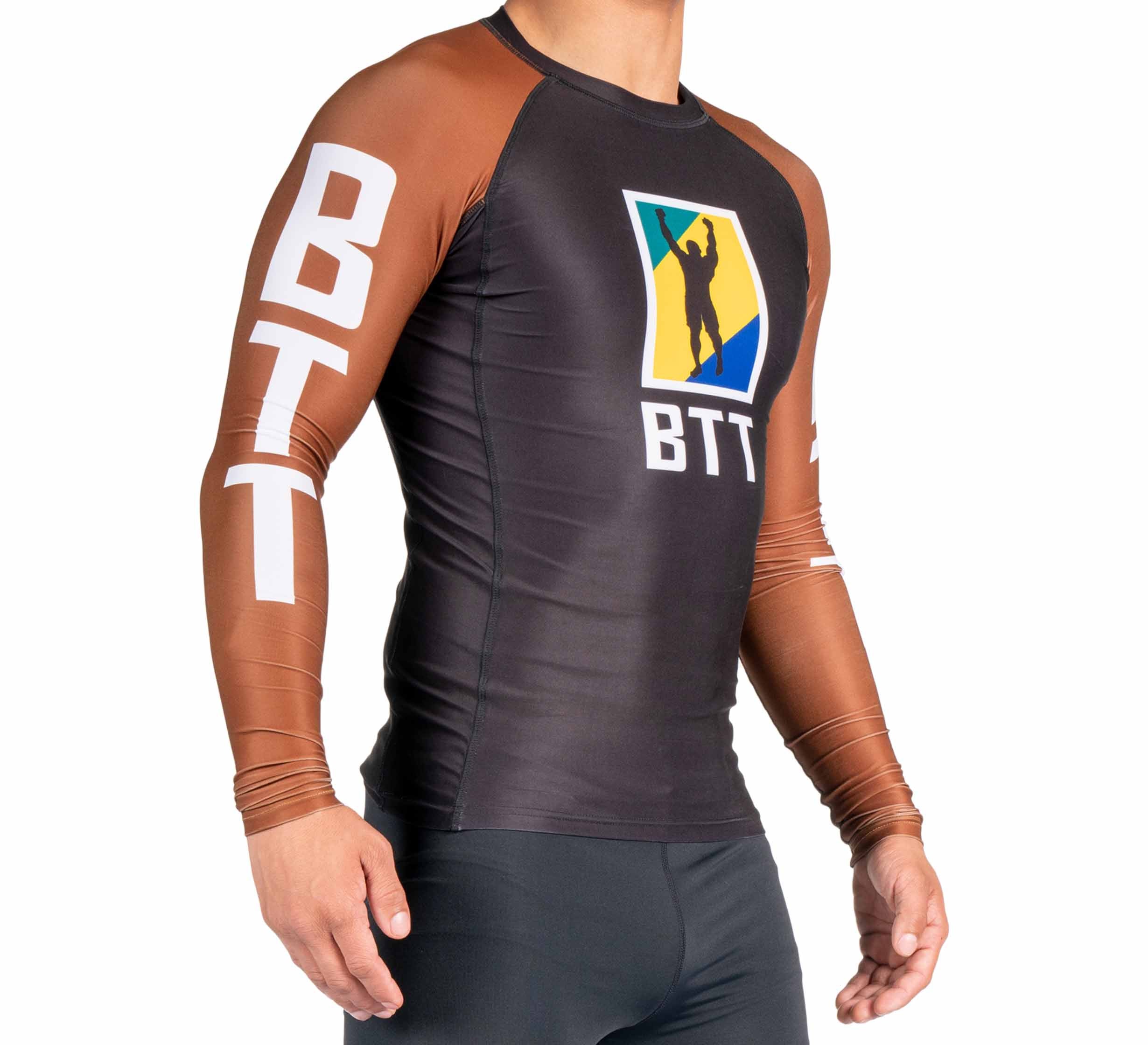 A person wears the BTT Ranked Long Sleeve Rashguard (NEW) featuring “BTT” on the brown sleeves, a colorful logo with a silhouette centered on the black chest.