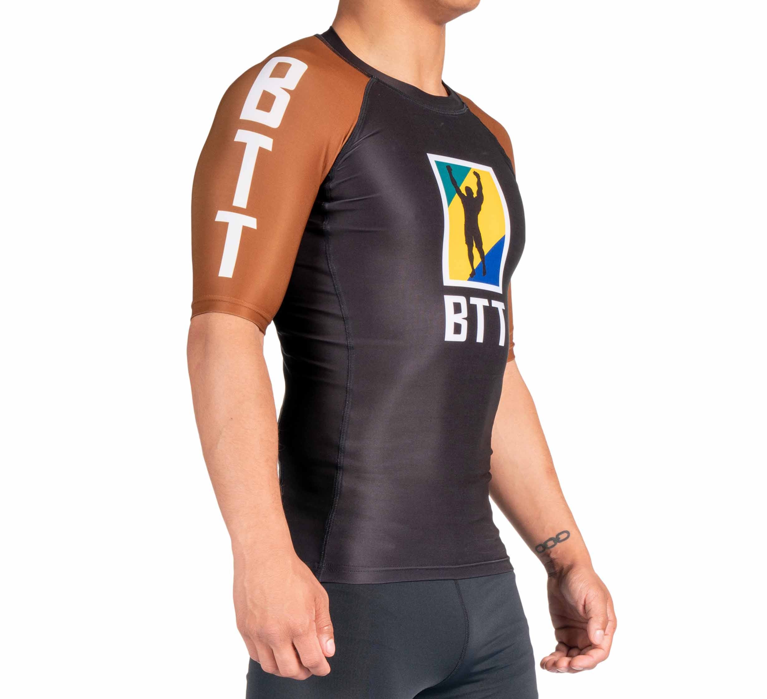 A person wears the BTT Ranked Short Sleeve Rashguard (NEW), a fitted black and brown top with "BTT" on the sleeves and a colorful chest logo featuring a silhouette, standing against a plain white background.