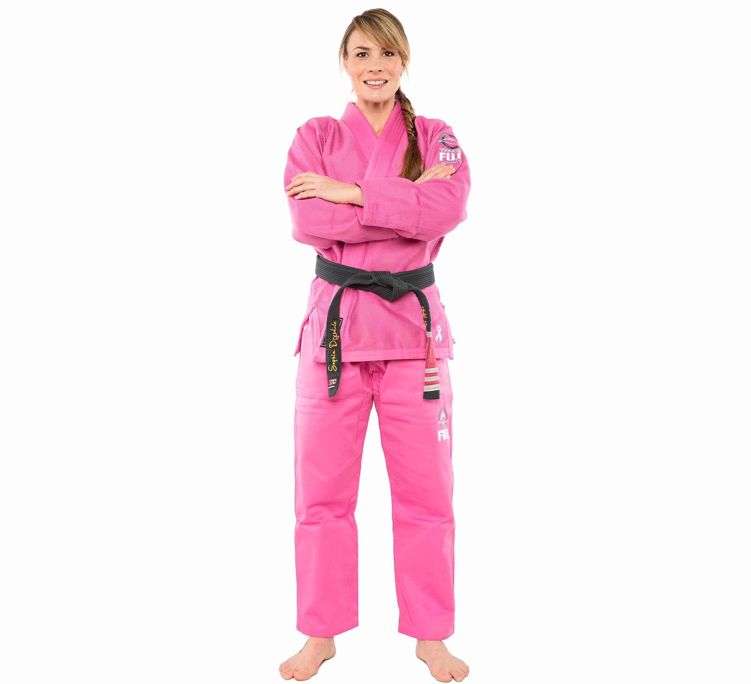 A woman stands barefoot, smiling with arms crossed, wearing the All Around Womens BJJ Gi Pink and a black belt against a plain white background.