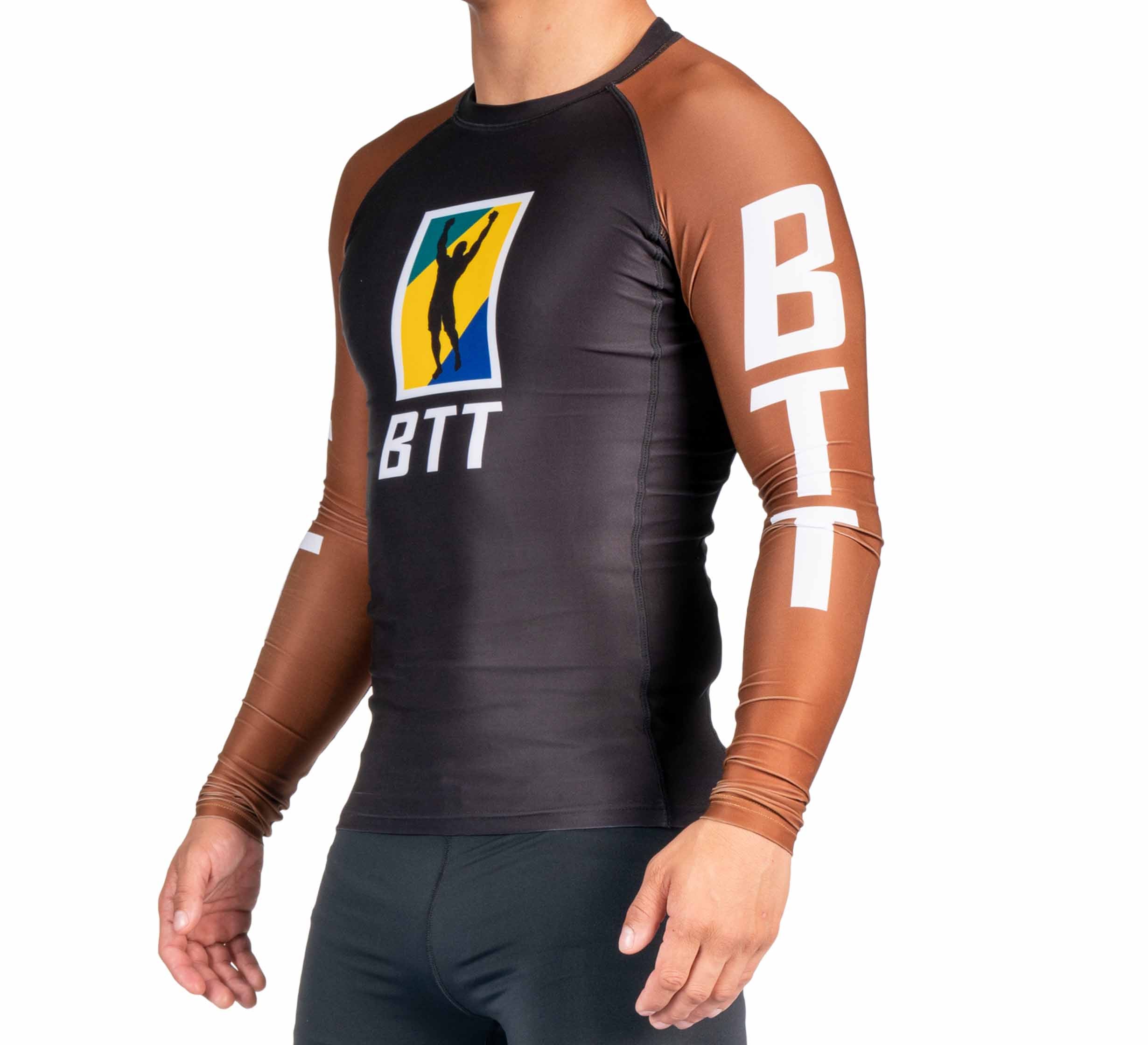 A man wears the BTT Ranked Long Sleeve Rashguard (NEW), a fitted top with brown sleeves, a black front, "BTT" lettering, and a graphic of a person raising an arm on the chest and sleeves.