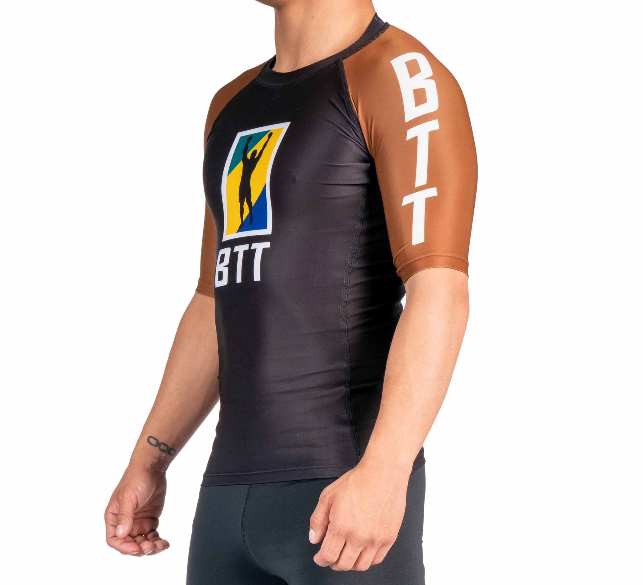 A person in the BTT Ranked Short Sleeve Rashguard (NEW), featuring black and brown colors, short sleeves, "BTT" text on the sleeve and chest, and a colorful central logo with a silhouette.