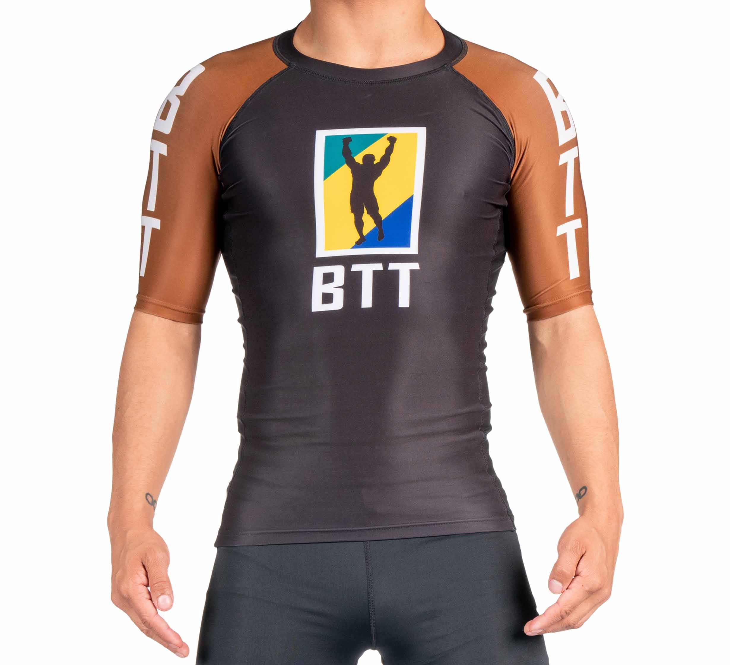 Someone wears the BTT Ranked Short Sleeve Rashguard (NEW), a black and brown athletic shirt with "BTT" and a silhouette logo of a person with raised arms on the chest, plus "BTT" on the sleeves.