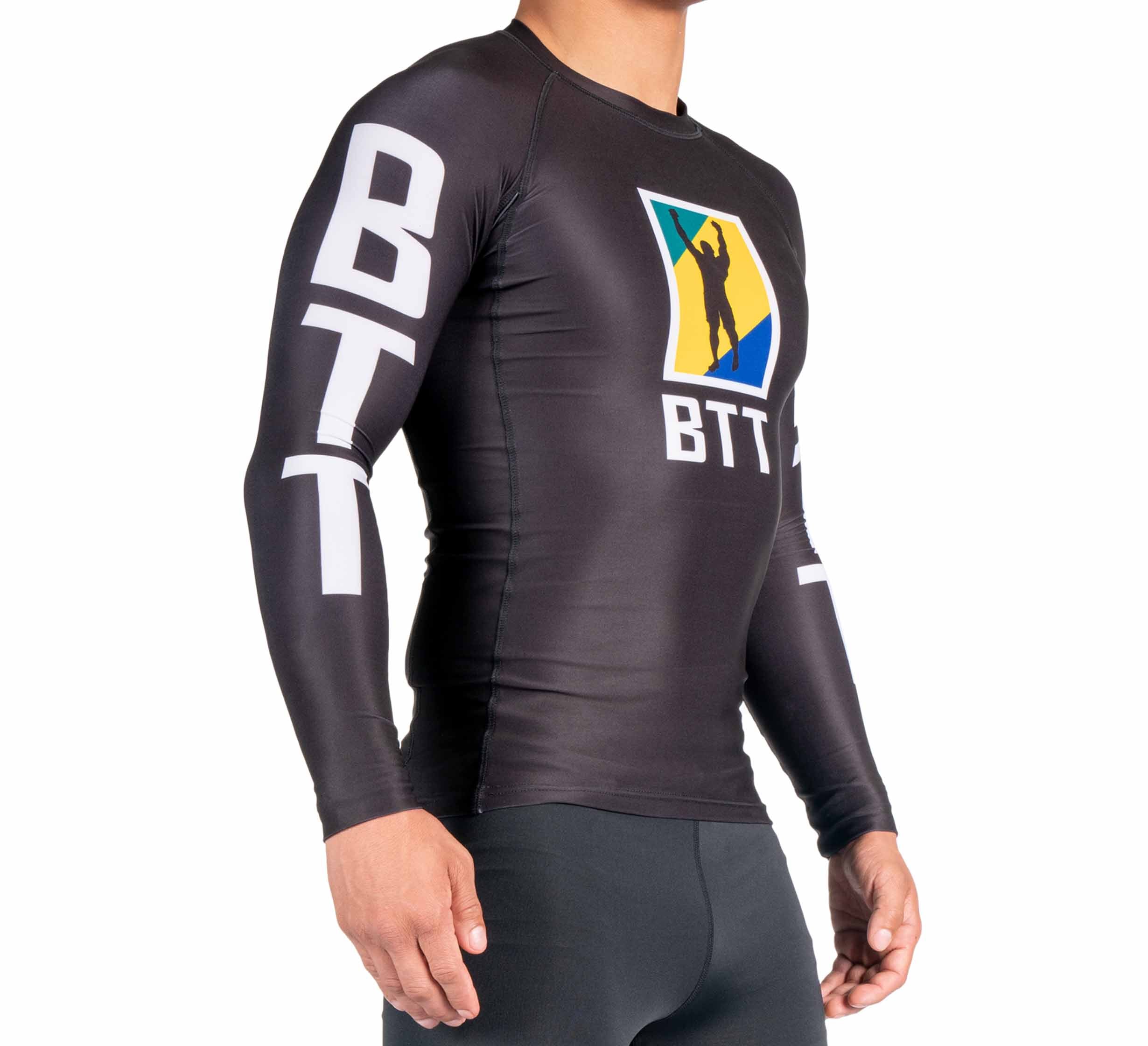 A person wearing the BTT Ranked Long Sleeve Rashguard (NEW) in black, featuring BTT lettering on the arms and a colorful chest logo with a silhouette figure, stands against a plain white background.