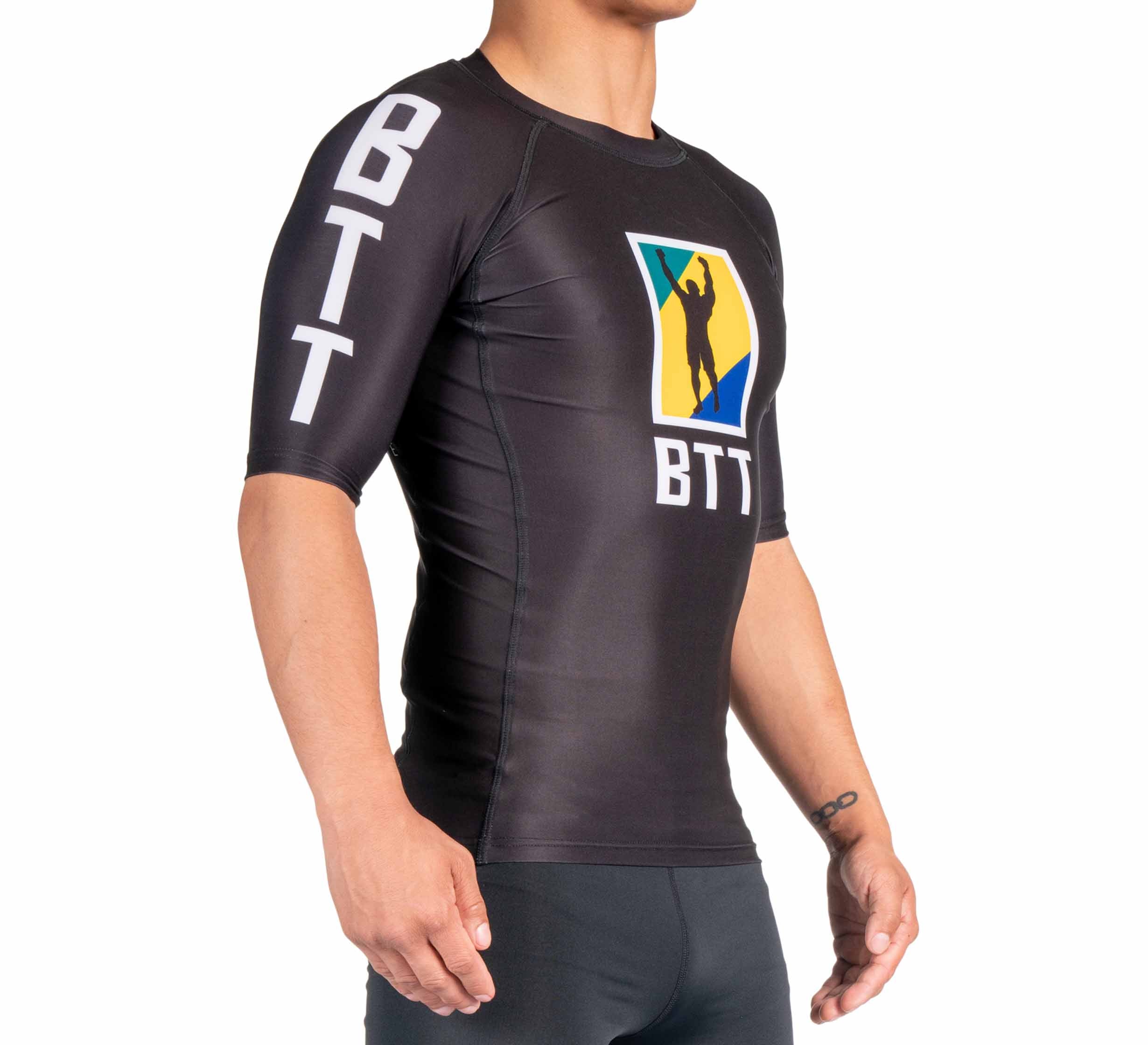 A person wearing the BTT Ranked Short Sleeve Rashguard (NEW), a tight black rash guard with BTT on the sleeves and a colorful logo featuring a person’s silhouette on the chest. The side photo highlights the upper body and design details.