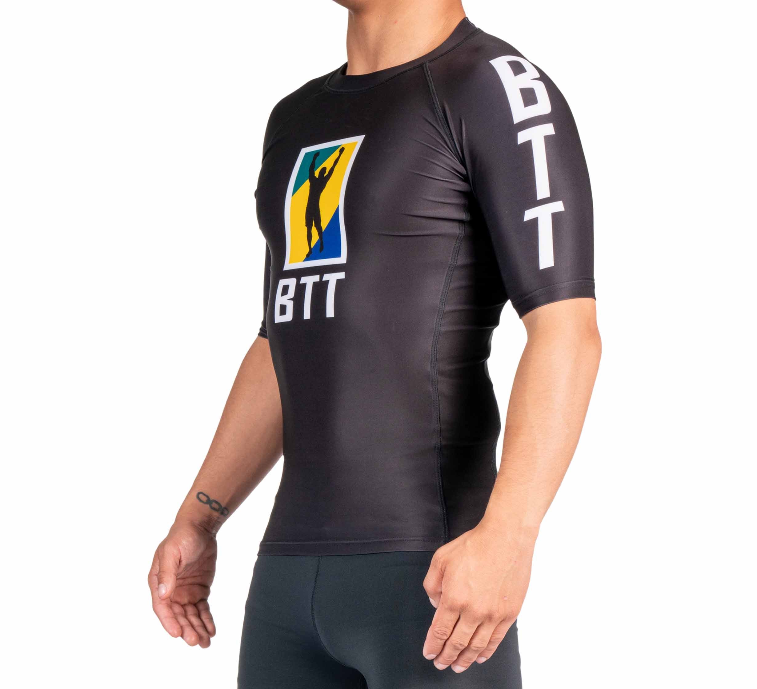 A person wearing the BTT Ranked Short Sleeve Rashguard (NEW), a fitted black athletic top with BTT on the sleeve and chest and a colorful silhouette logo, stands against a white background.