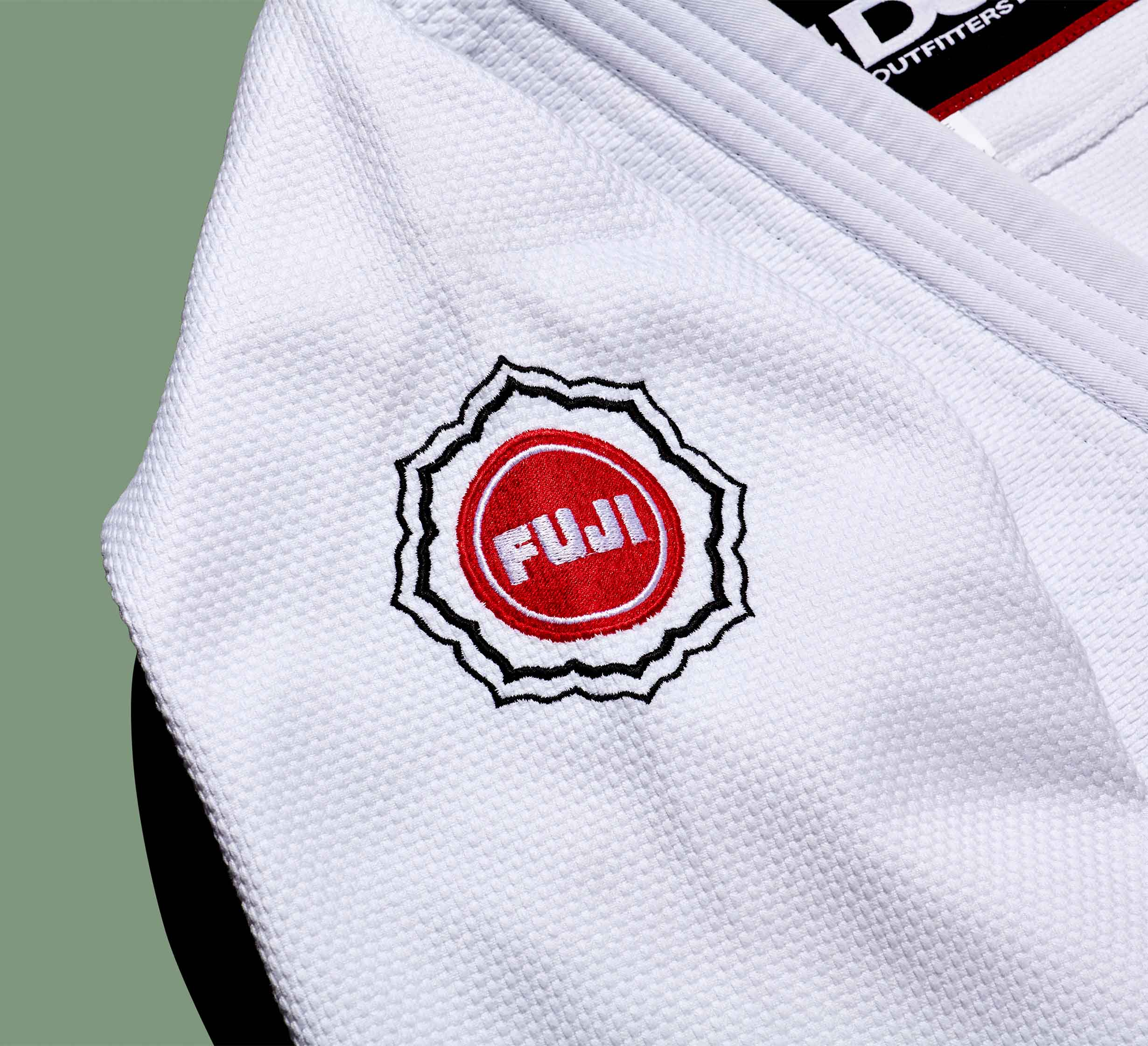 Close-up of the Dojo Outfitters x FUJI Judo Gi, featuring textured white fabric and a red and white Fuji logo patch on the shoulder, set against a solid muted green background.