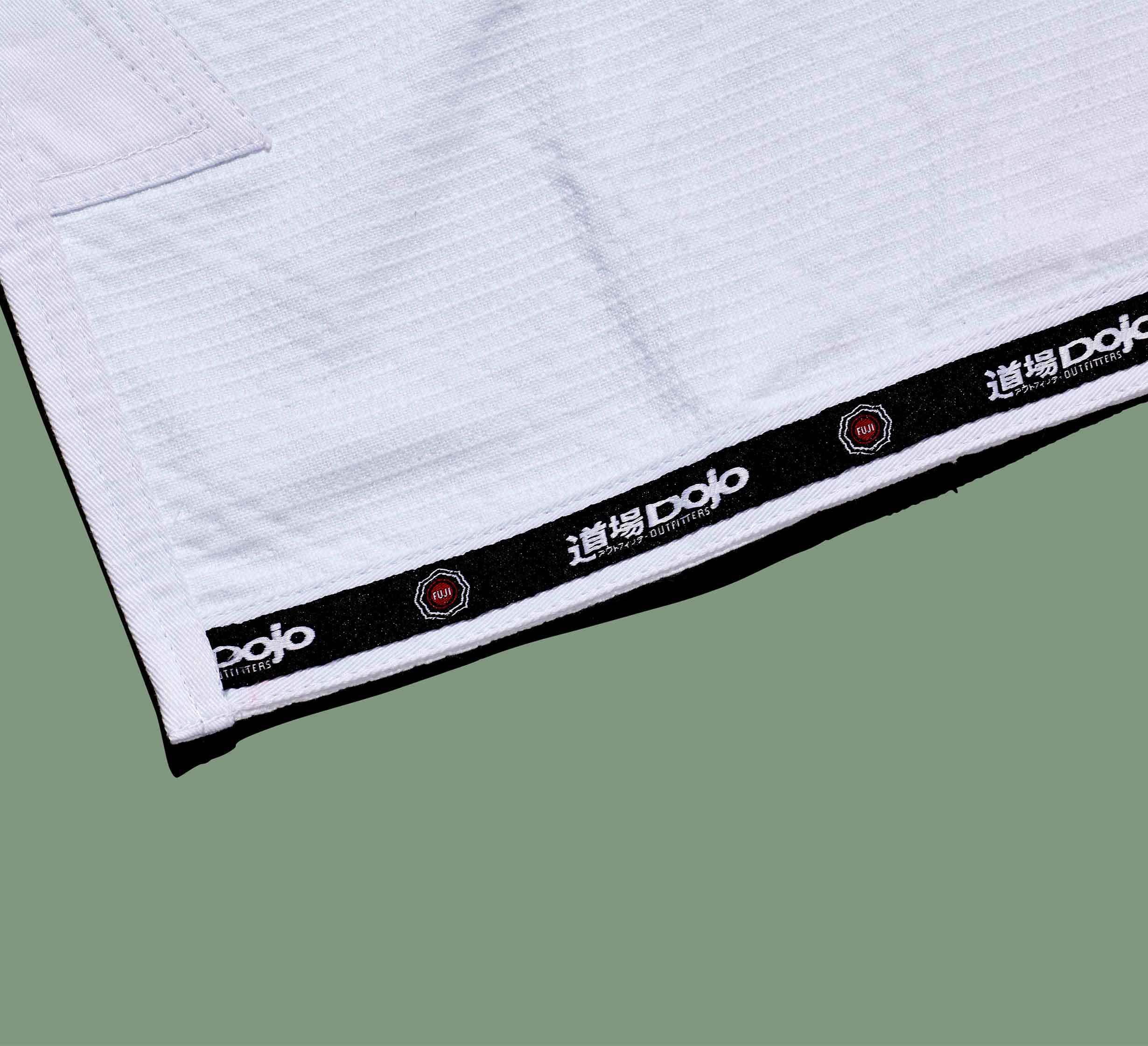Close-up of the hem of a Dojo Outfitters x FUJI Judo Gi, white with black trim, white Japanese characters, "Dojo" text, and small red octagonal logos on a sage green background.