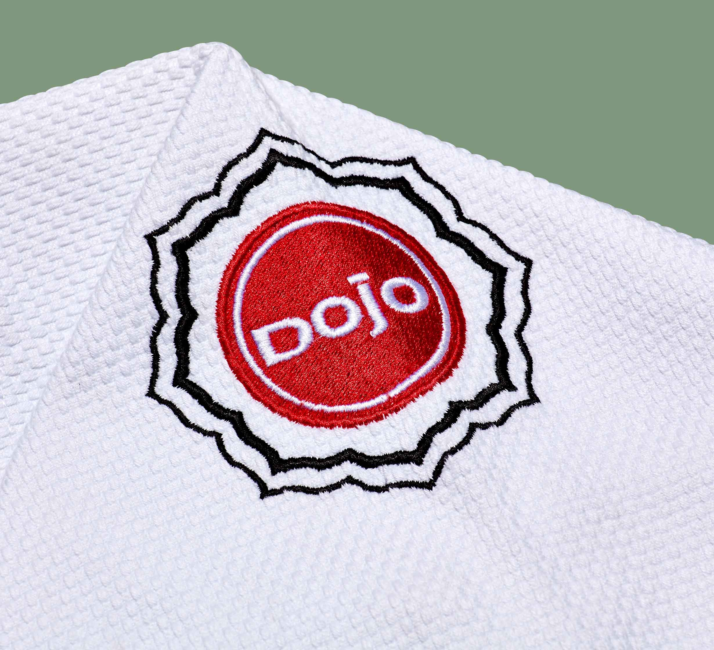 A close-up of the Dojo Outfitters x FUJI Judo Gi, featuring a white textured fabric and a red and white circular dojo patch outlined in black stitching on the shoulder.