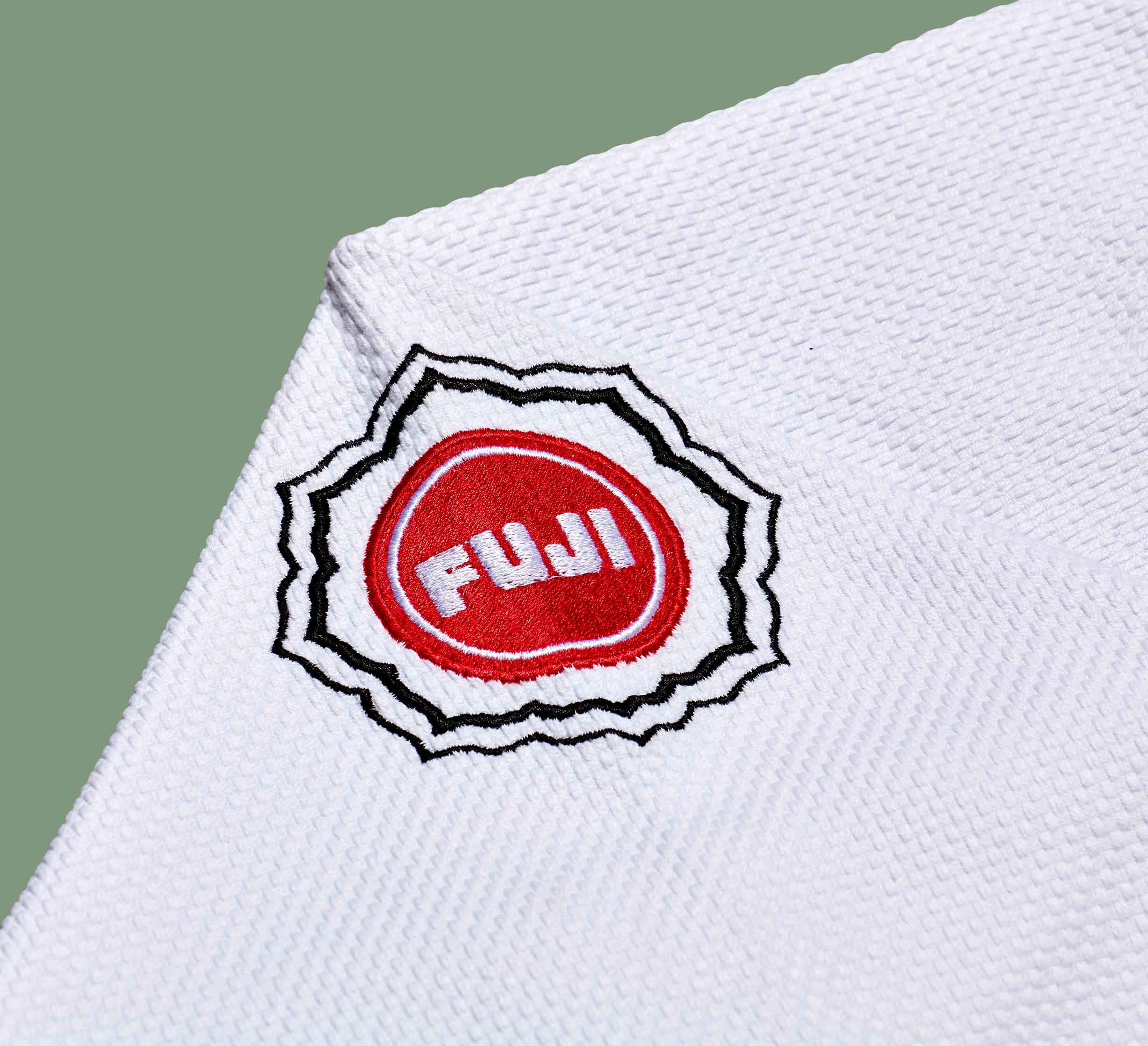 Close-up of a white textured Dojo Outfitters x FUJI Judo Gi fabric with a red and white Fuji logo patch embroidered, set against a green background.
