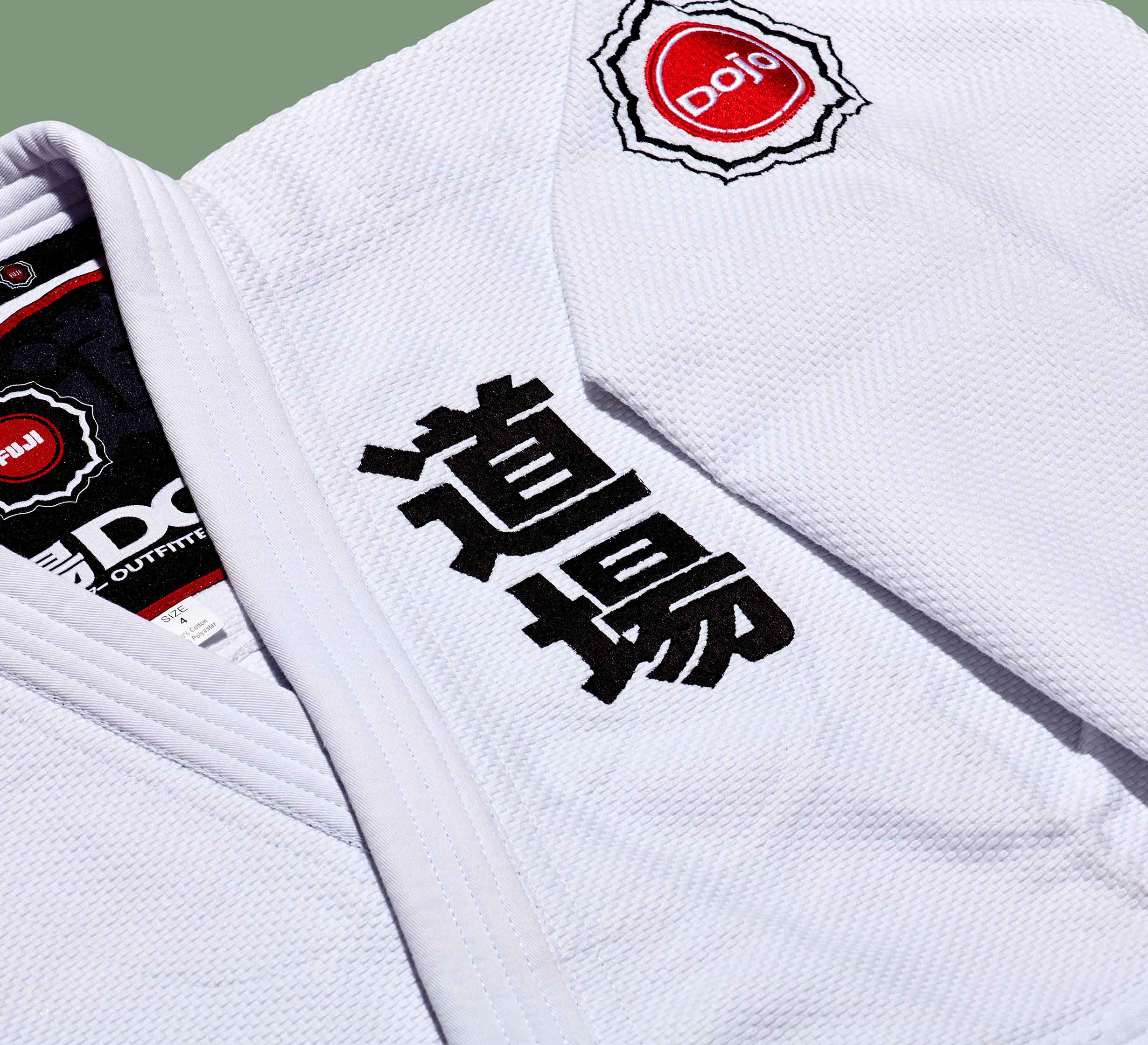 A close-up of the Dojo Outfitters x FUJI Judo Gi shows sturdy, textured white fabric, black Japanese kanji characters, and a red circular “dojo” patch on the shoulder.