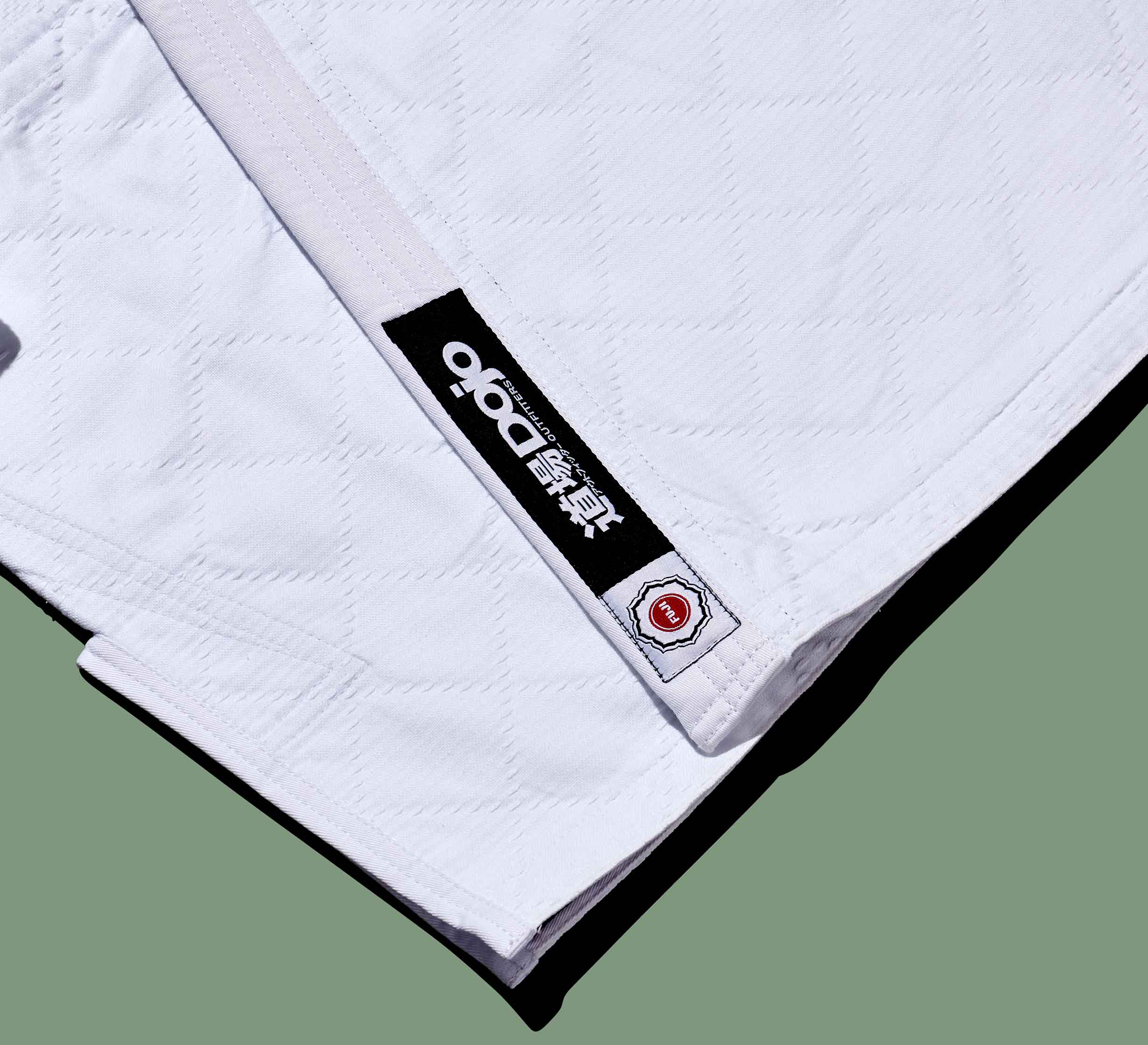 Close-up of the Dojo Outfitters x FUJI Judo Gi featuring a white quilted texture, a black and white DRO patch, Japanese kanji, and a small red and white embroidered patch near the hem.