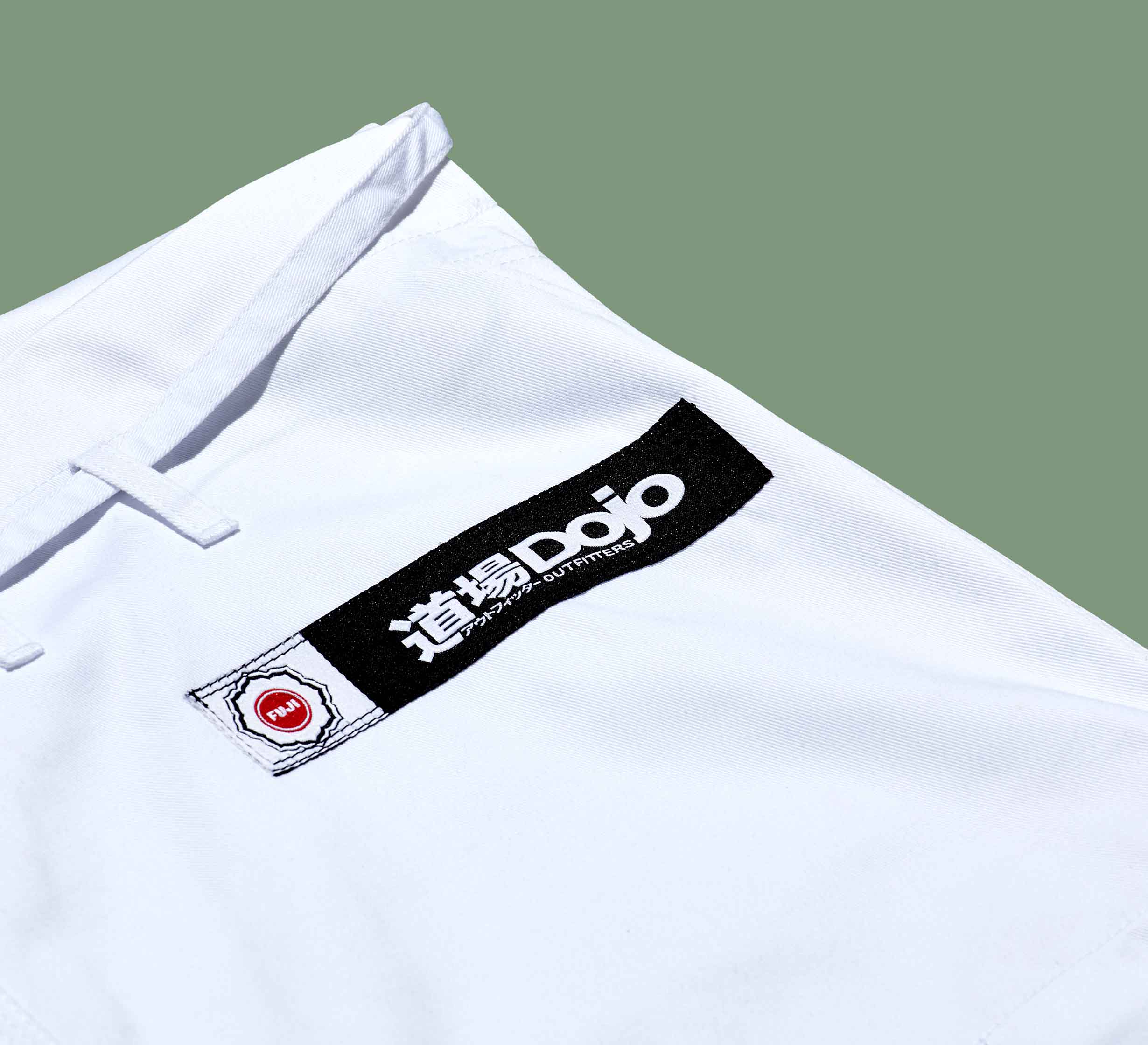 Close-up of the Dojo Outfitters x FUJI Judo Gi, a white martial arts uniform with a black patch displaying "Dojo" and Japanese characters, plus a smaller white patch with a red circle, set against a light green background.