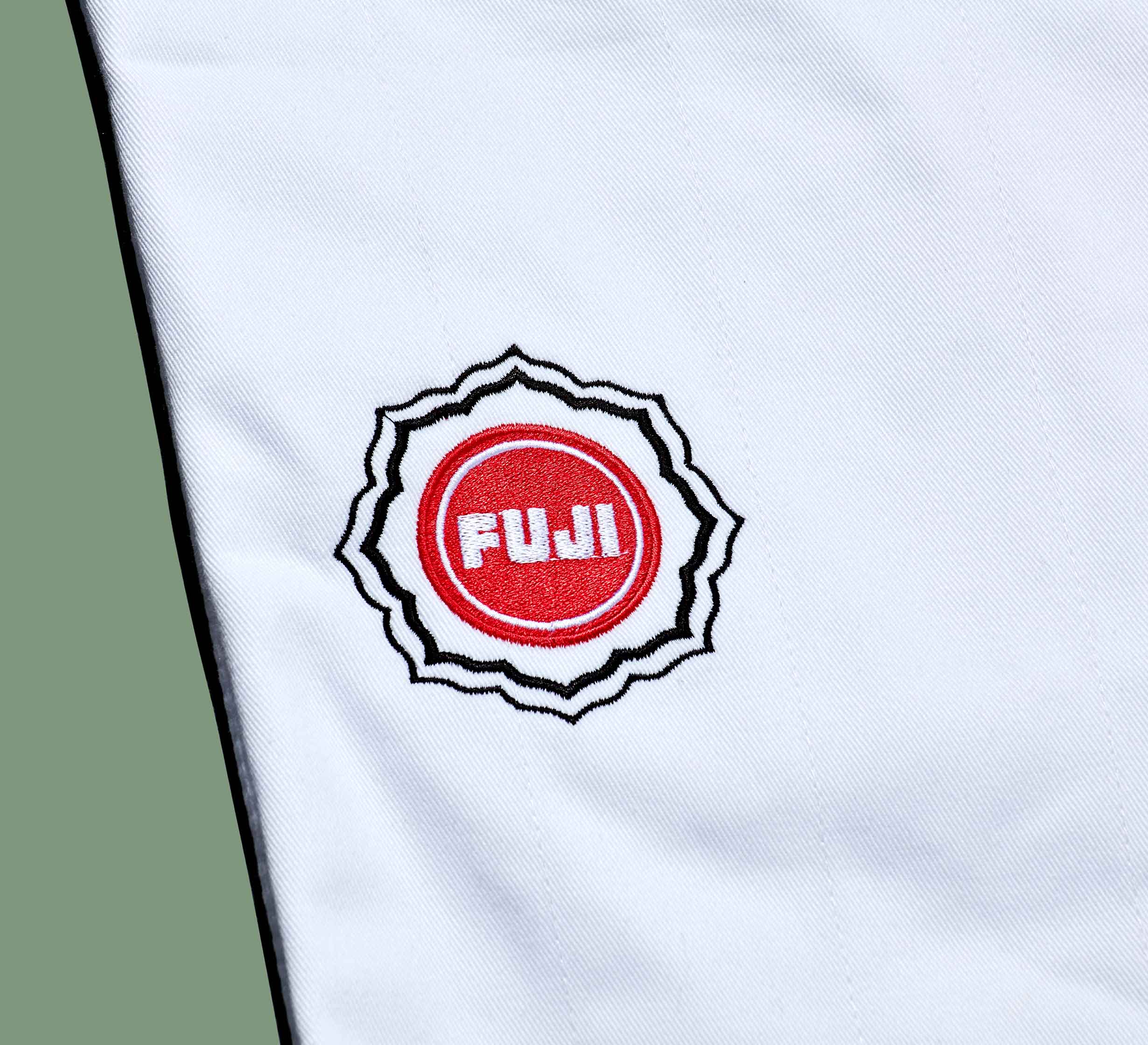A white Dojo Outfitters x FUJI Judo Gi features a red circular FUJI logo, outlined in black and white like a badge or patch, stitched onto the fabric. The background is light green.