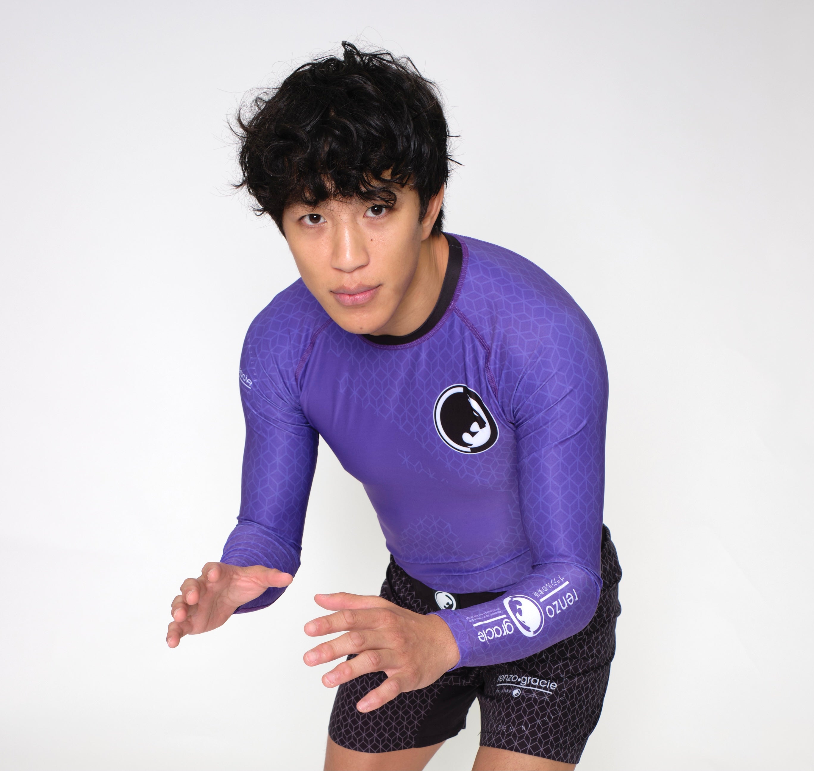 A person with short, curly dark hair wears a purple Renzo Gracie Mecca Ranked Long Sleeve Rashguard and black shorts, standing in a grappling stance against a plain white background.