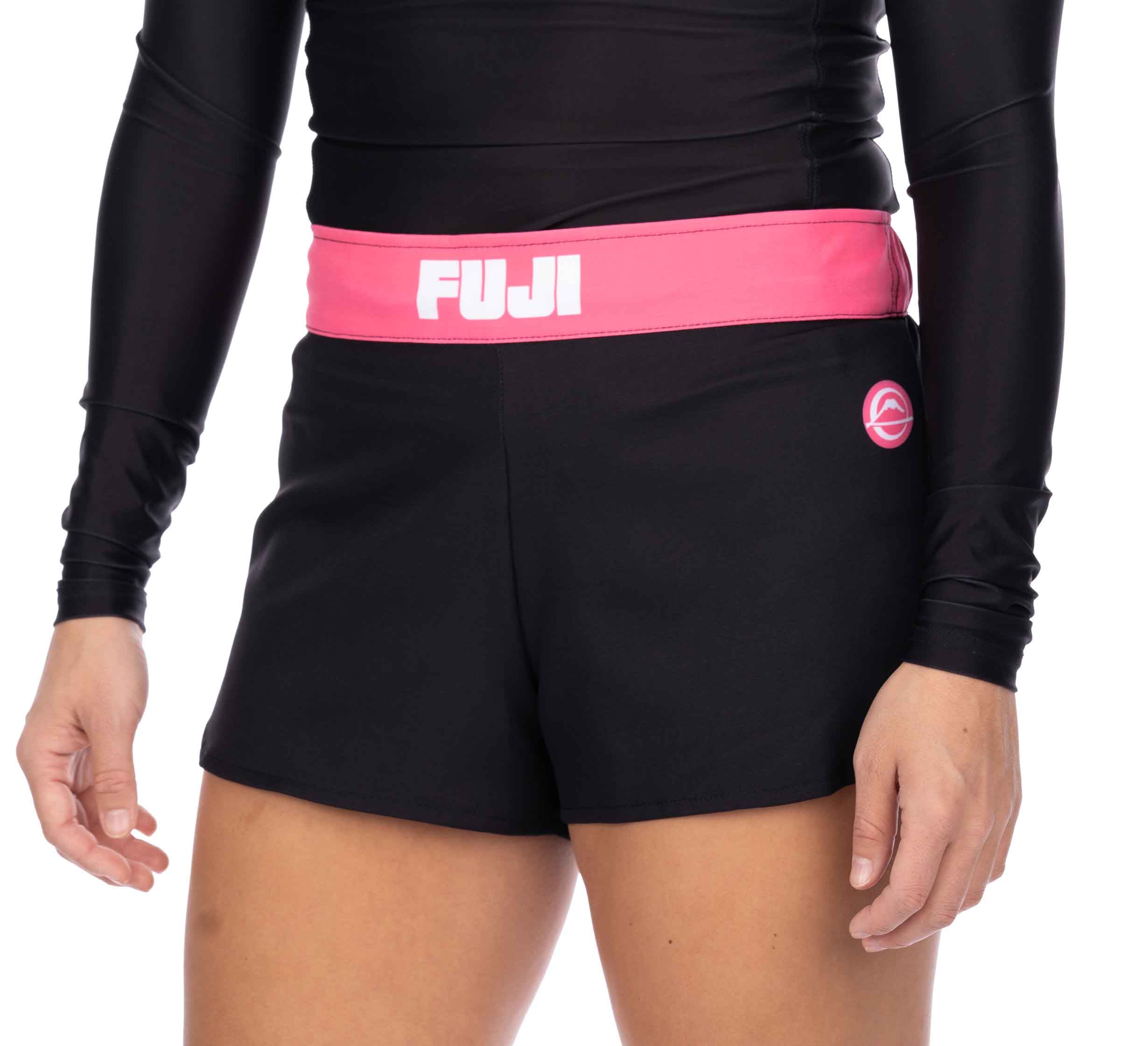 A person wearing Essential Grappling Pink Fight Shorts, featuring a black design with a pink waistband labeled FUJI and a matching pink logo on the left leg, paired with a black long-sleeve top.