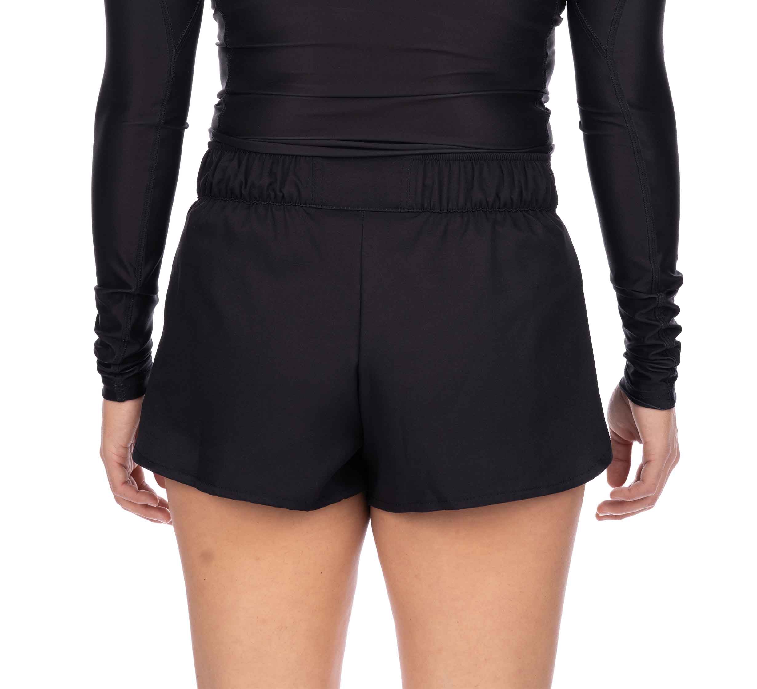 A person is shown from the back wearing Essential Grappling Black Fight Shorts with an elastic waistband and a long-sleeve black top, arms relaxed at their sides.