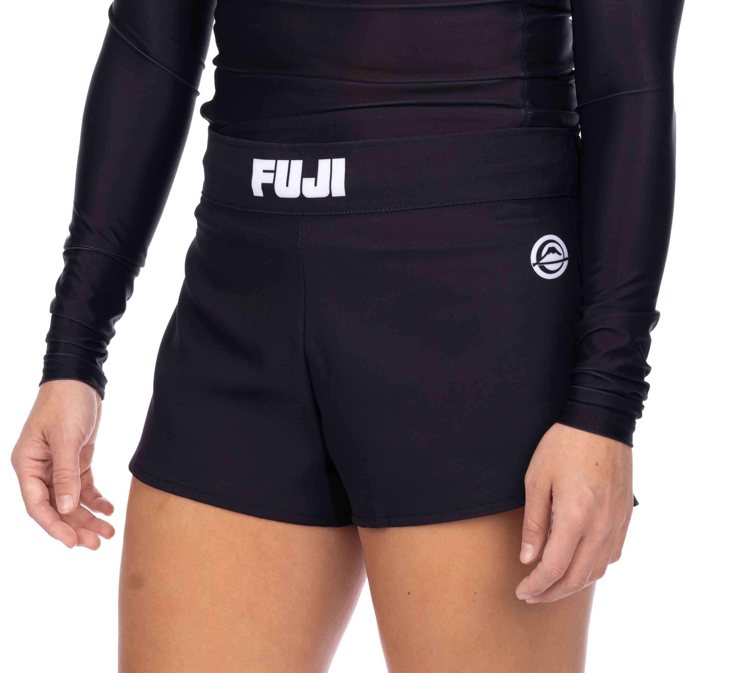 A person wearing Essential Grappling Black Fight Shorts with "FUJI" on the waistband and a circular logo on the left leg, paired with a black long-sleeve top. The image highlights the shorts and lower torso.