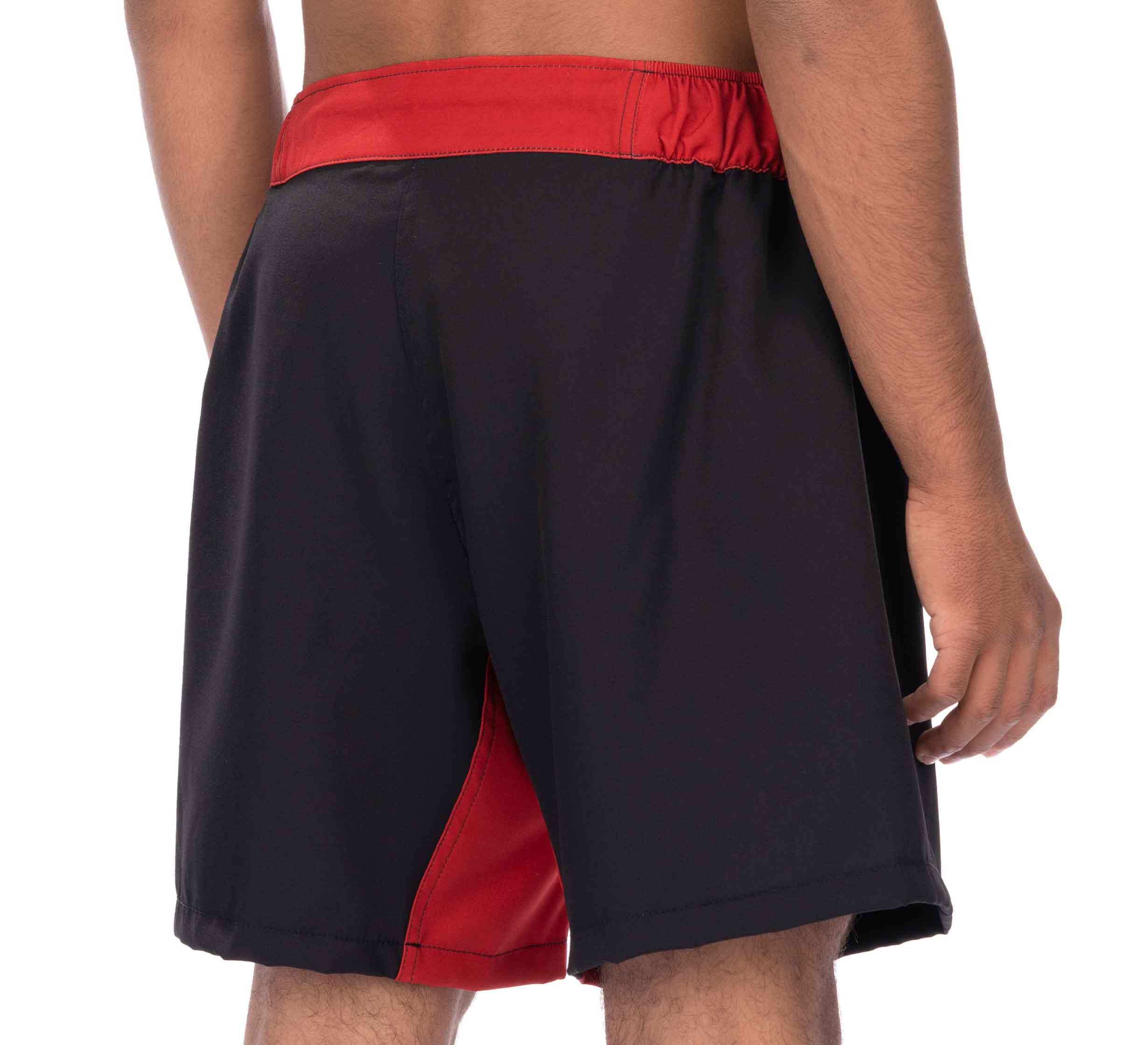 A person wearing Essential Grappling Red Fight Shorts, featuring a black body with a red waistband and red side panel, is photographed from the back with their right arm relaxed by their side.