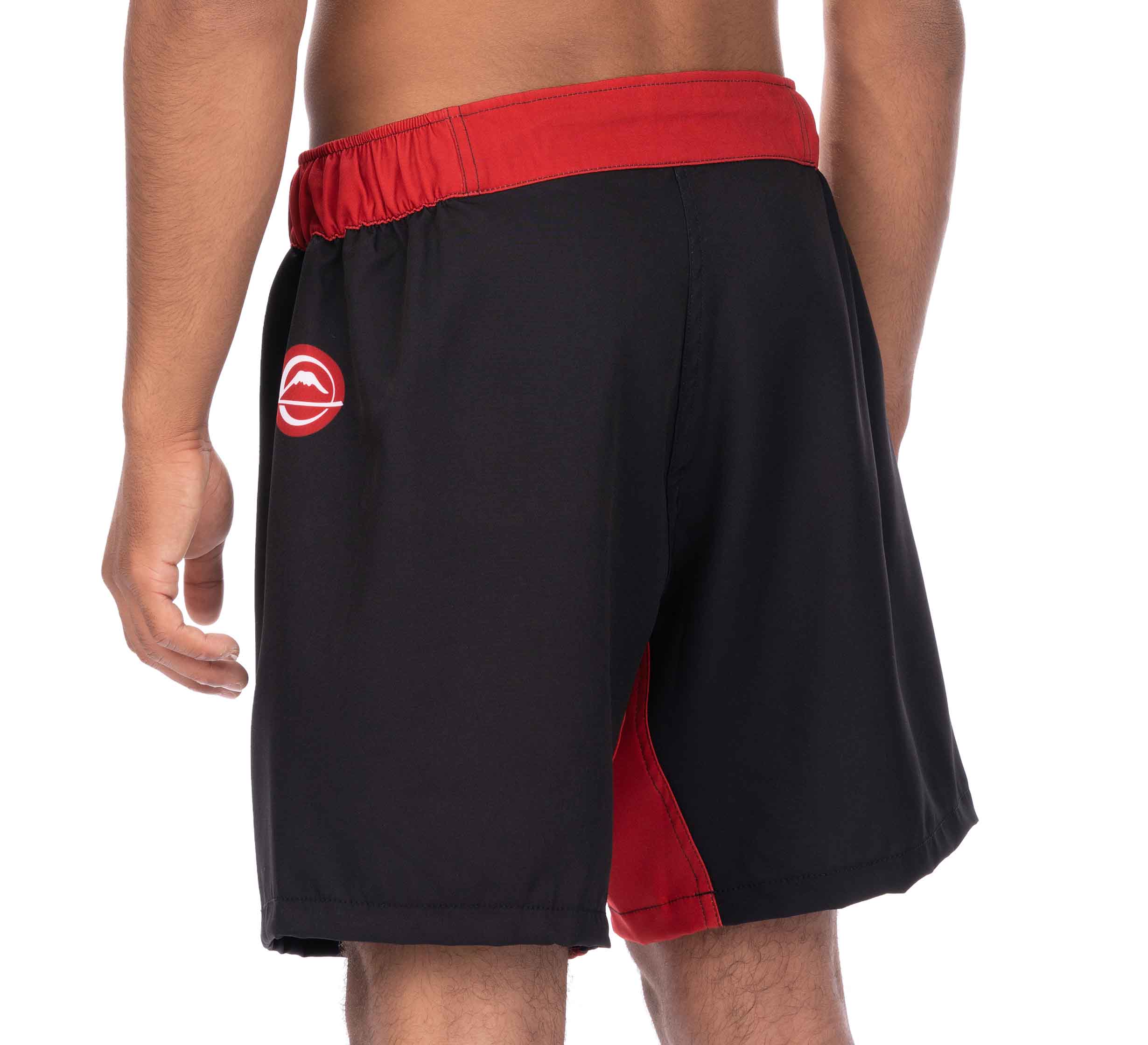 A person wearing Essential Grappling Red Fight Shorts, black with a red waistband and side accents, viewed from the back. The left side of the shorts features a white logo.