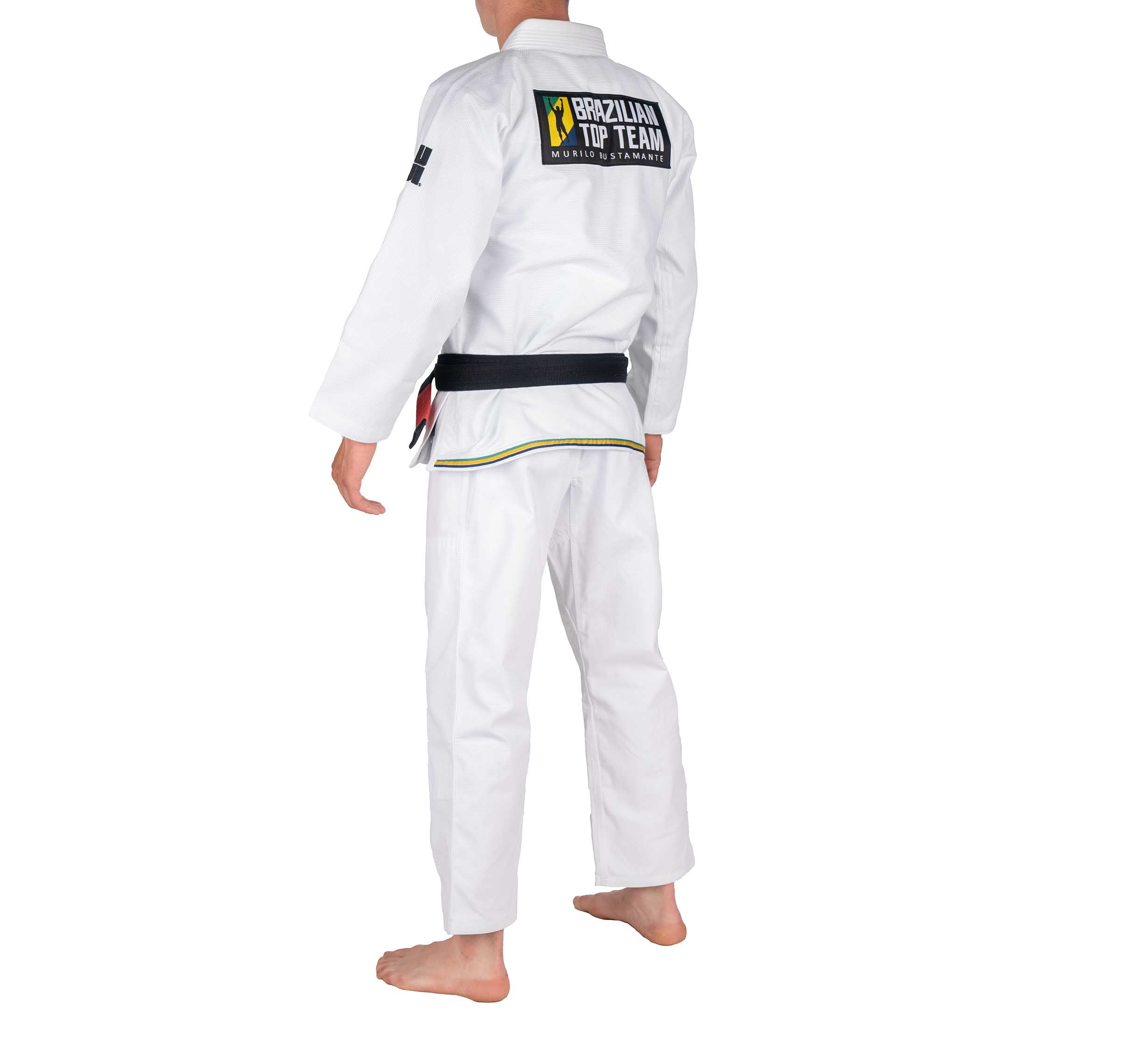 A person wearing the BTT Standard Gi - White with a black belt stands barefoot, facing away. The gi's back displays a Brazilian Top Team patch.