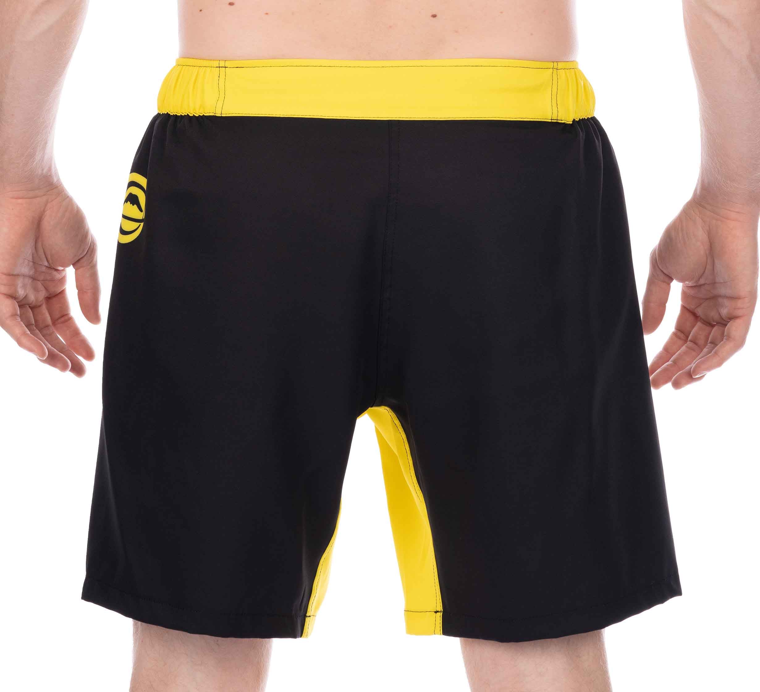 A person is shown from the waist down, facing away, wearing Essential Grappling Yellow Fight Shorts with a yellow waistband and a small logo on the left side. Their hands are relaxed at their sides.