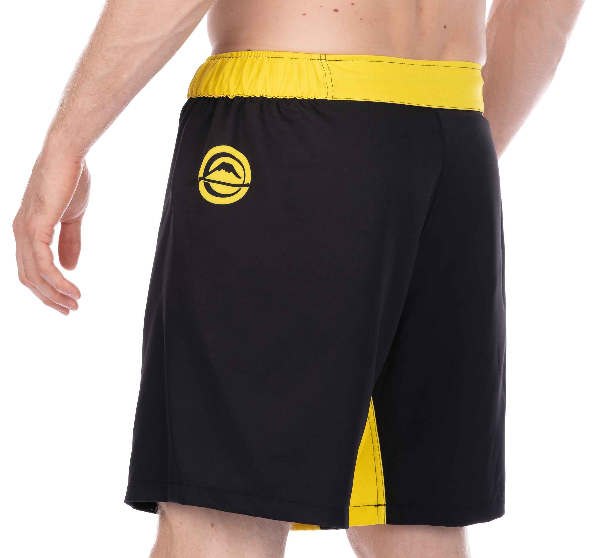 A person is wearing Essential Grappling Yellow Fight Shorts, which are black with a yellow waistband, side stripe, and a yellow circular logo on the left hip. Their back and left arm are visible.