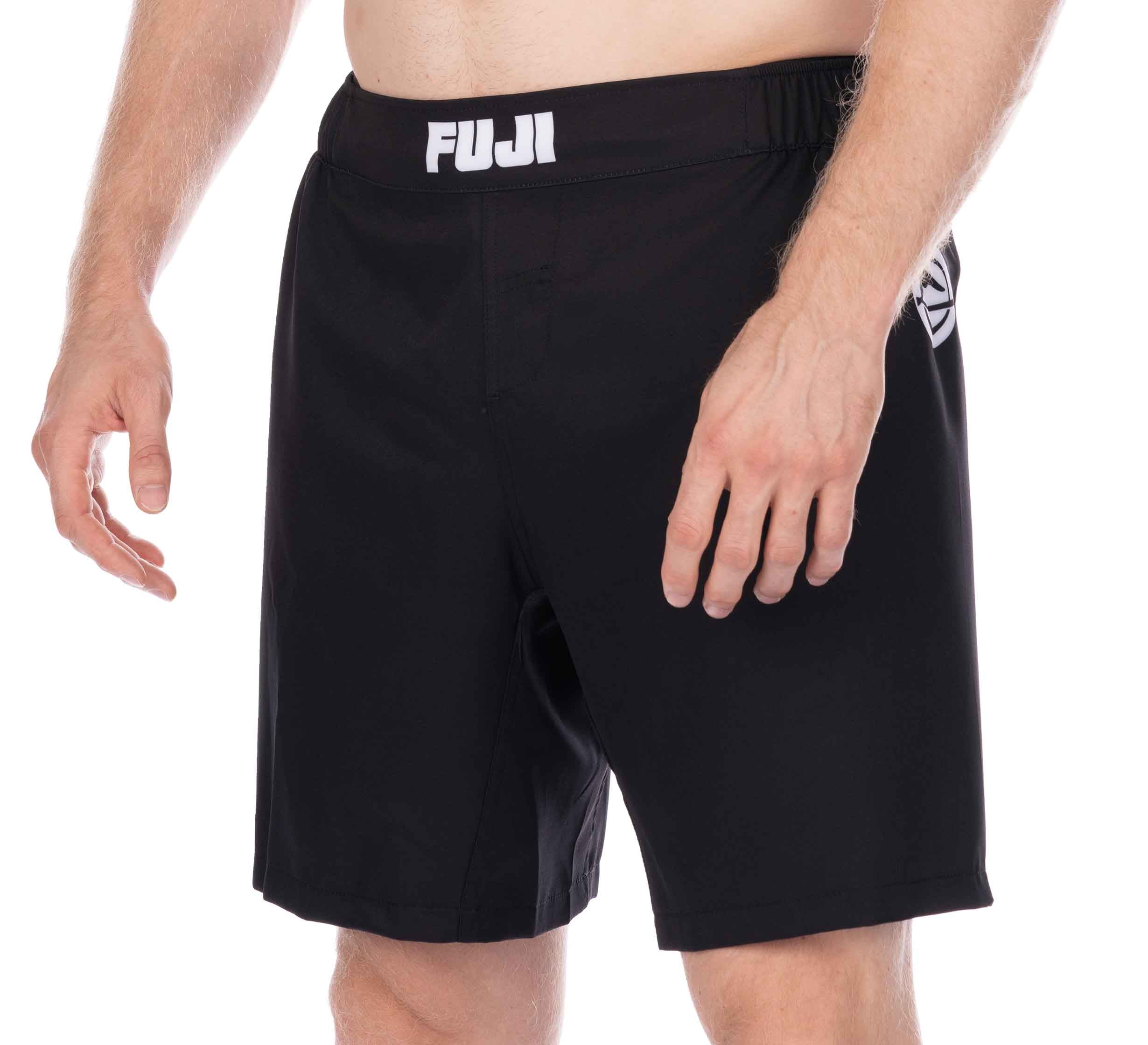A person wearing black Essential Grappling Black Fight Shorts, shown from waist to mid-thigh. The shorts feature a logo on the waistband and a small emblem on the left leg; their torso and arms are partially visible.