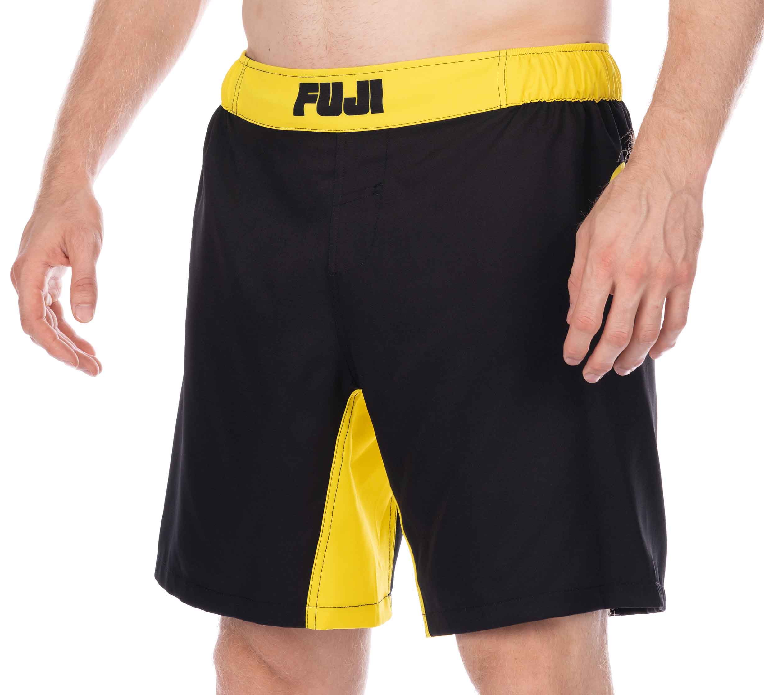 A person wearing Essential Grappling Yellow Fight Shorts with a black and yellow design and FUJI on the waistband, standing with hands relaxed by their sides.