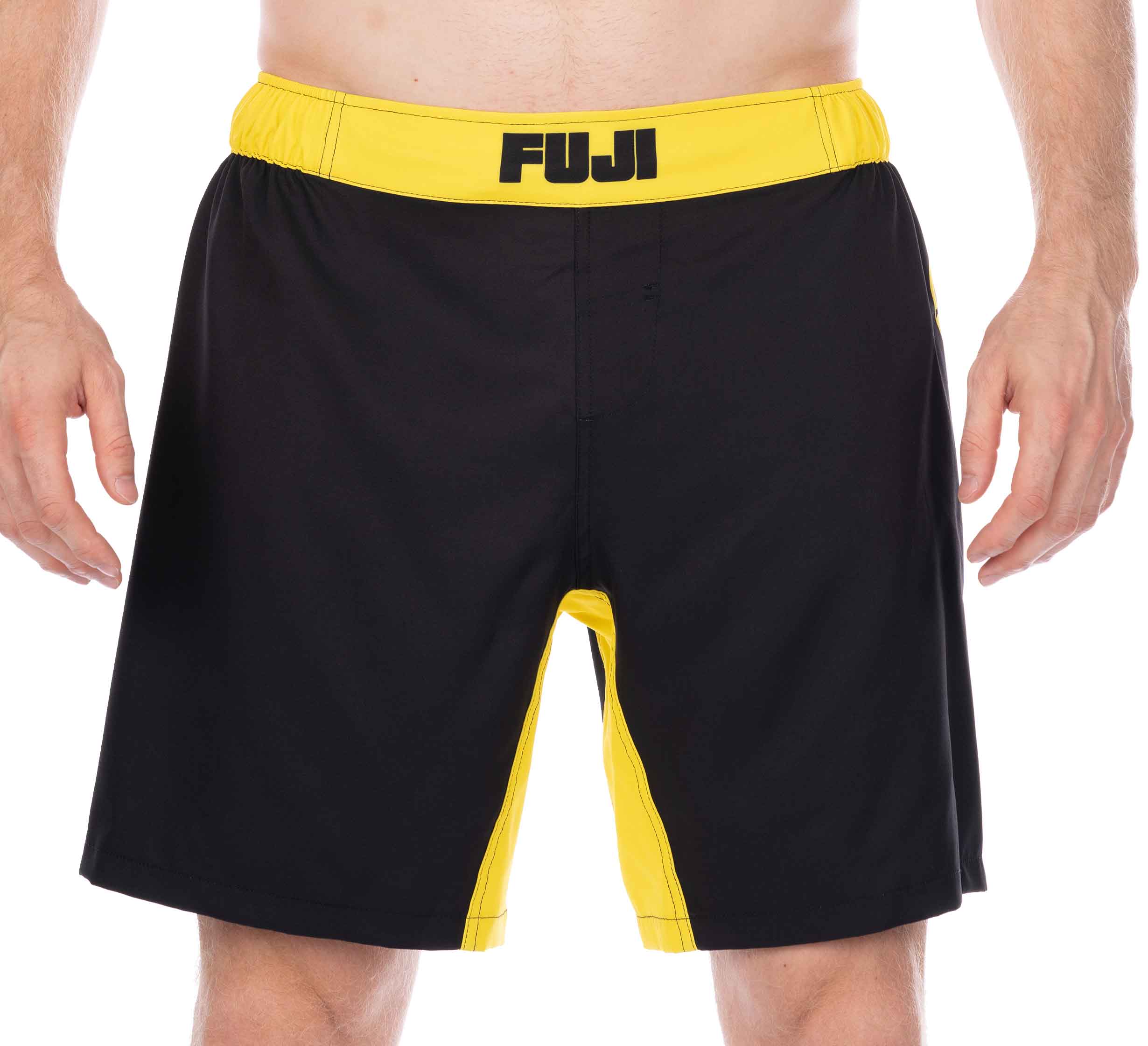 A person wearing Essential Grappling Yellow Fight Shorts, featuring black and yellow colors with the FUJI logo on the waistband, stands against a white background. Only their torso and upper legs are visible.