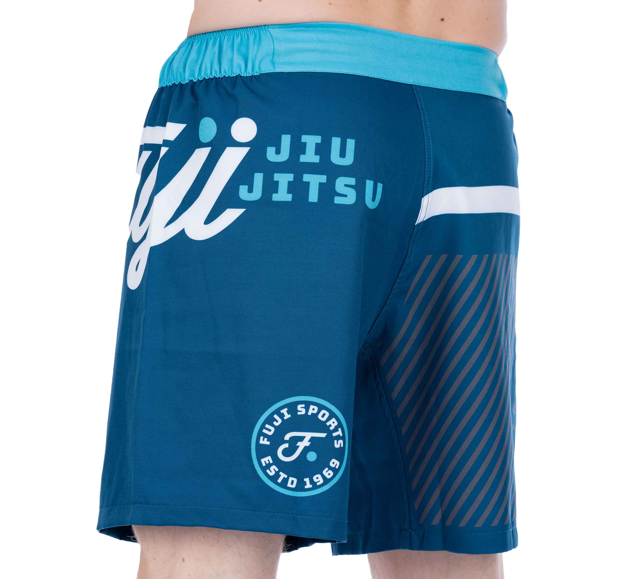 A person shown from the side and back is wearing Script Grappling Shorts Blue by Fuji Sports, featuring a turquoise waistband, white logo text, and diagonal brown side stripes.