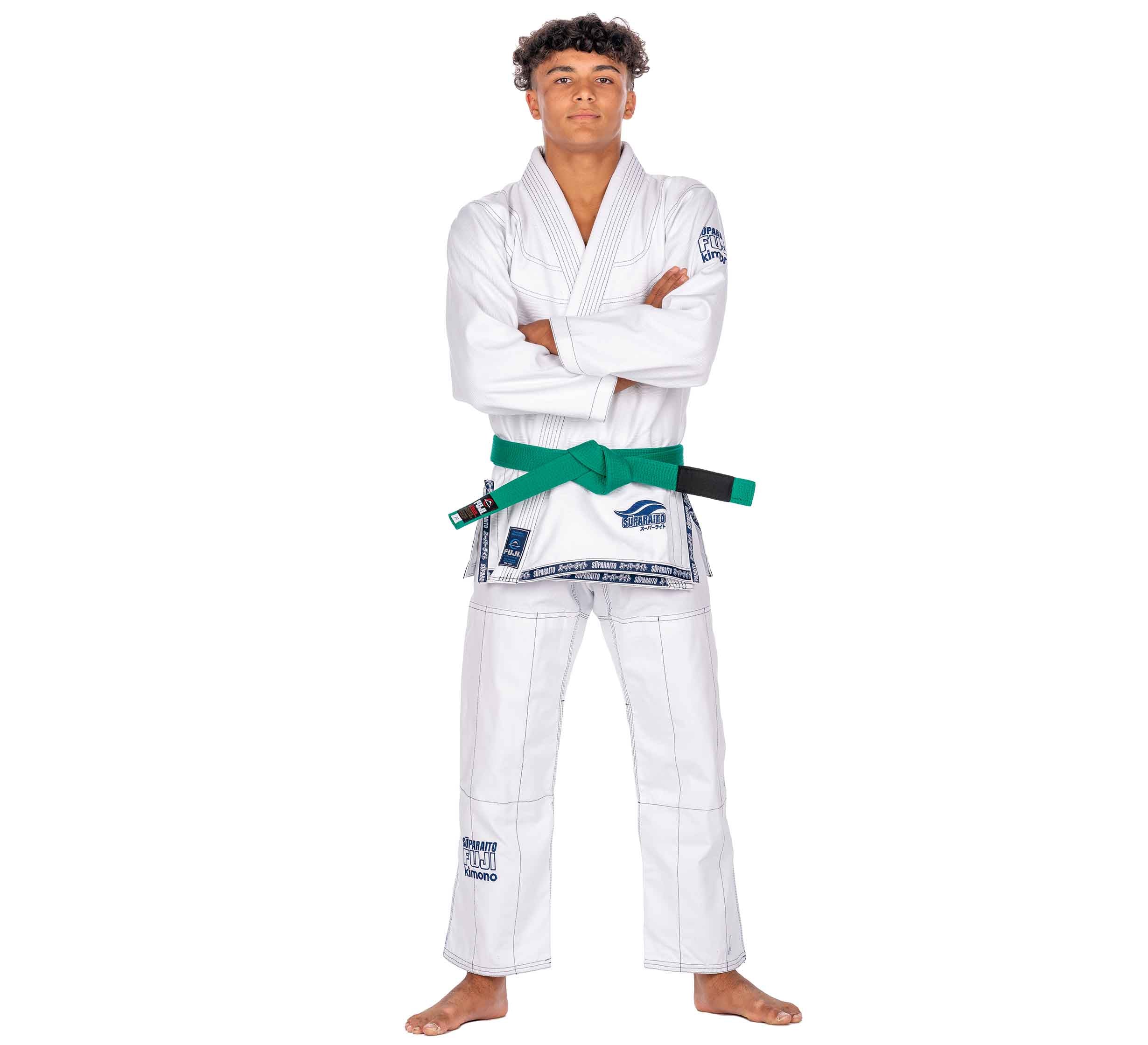 A young person stands barefoot with arms crossed, wearing the Suparaito Kids White BJJ Gi with a green belt. The gi features patches with blue text and logos on the chest, sleeve, and pants.