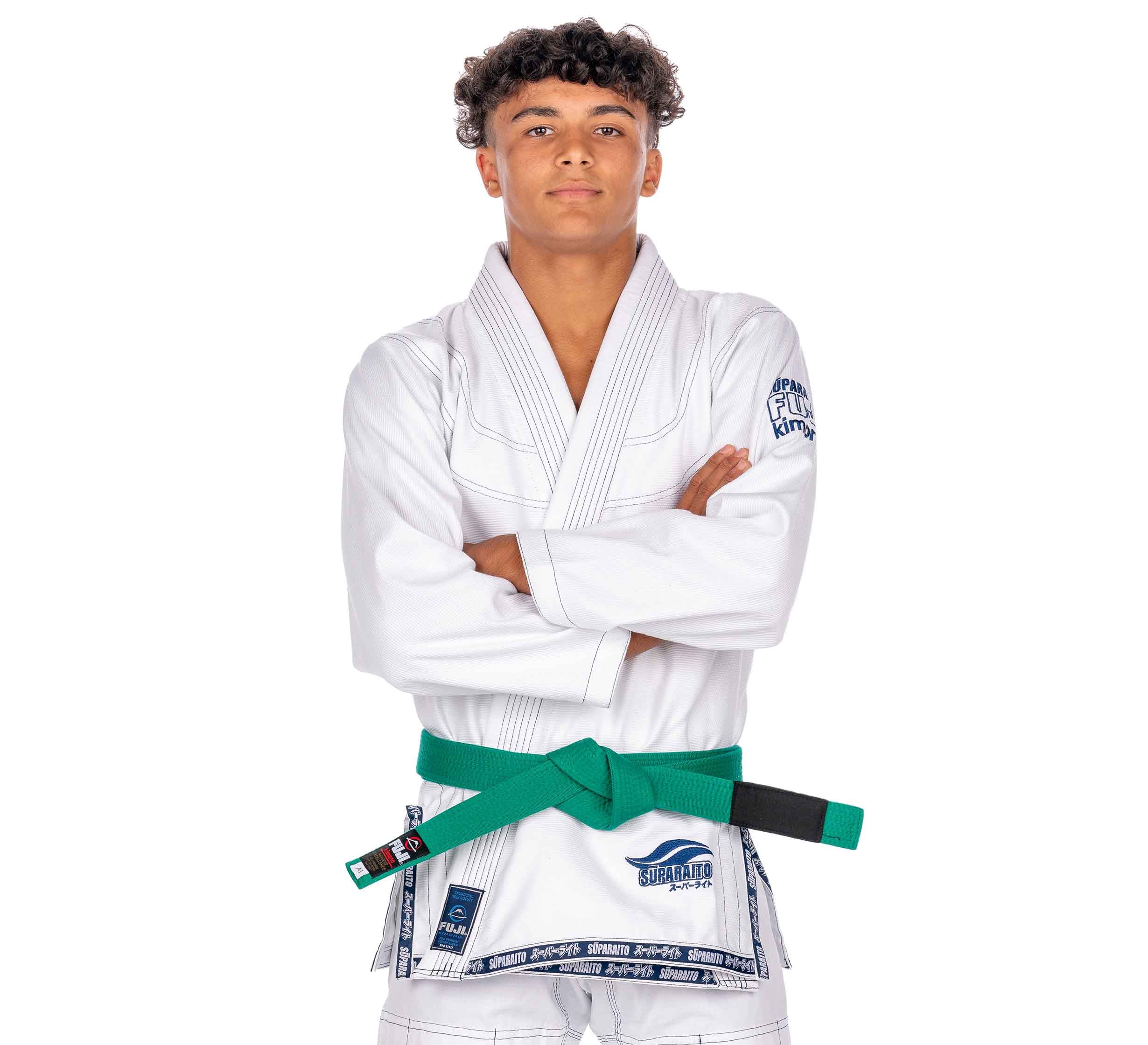 A confident young person with curly hair wears the Suparaito Kids White BJJ Gi and a green belt, standing with arms crossed against a white background.